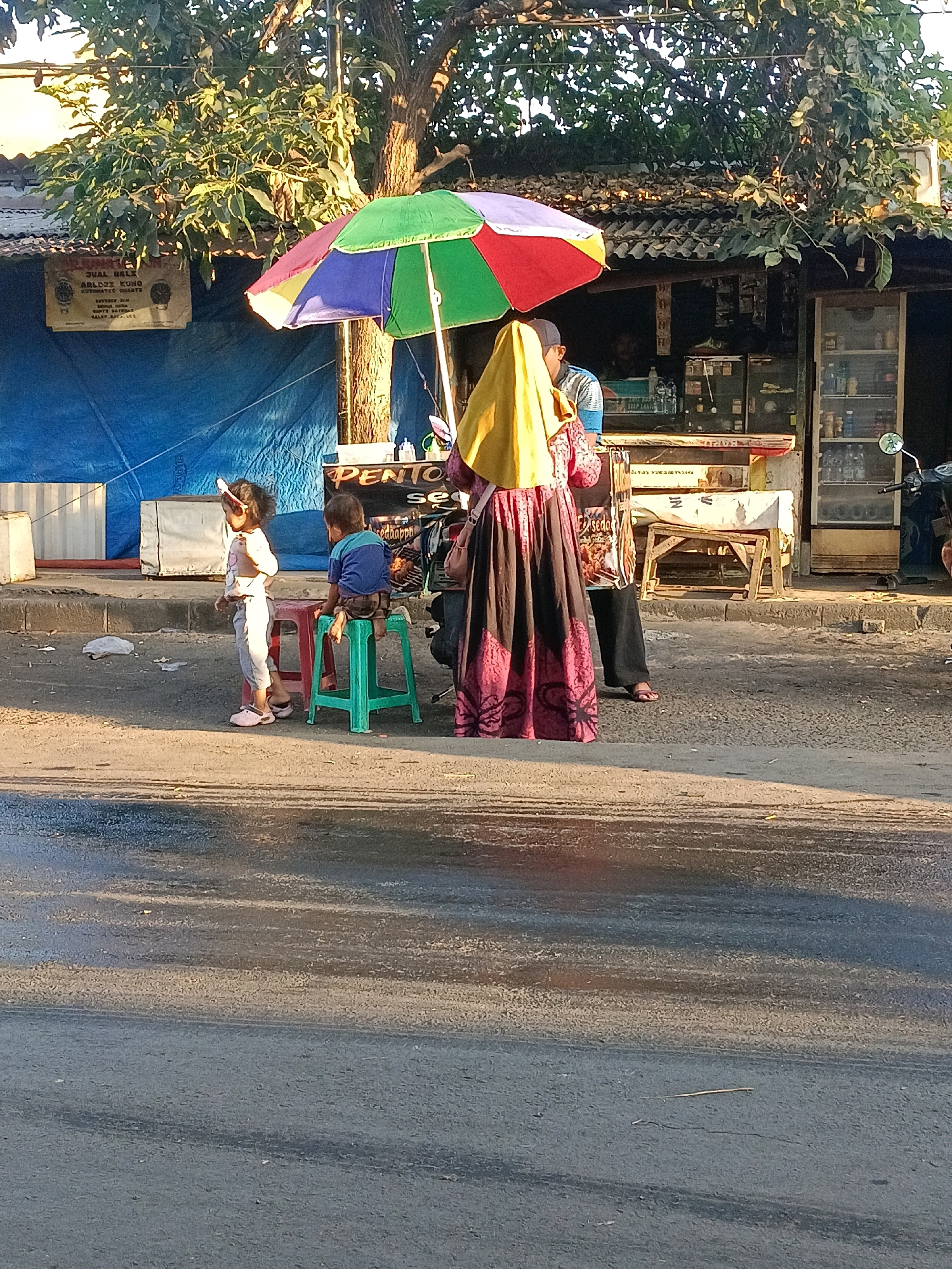 This is a medium-angle, full shot of a street scene in Wonocolo, Indonesia, during the day. The foreground shows a wet asphalt road reflecting the warm sunlight. In the middle ground, on the right side of the road, a small storefront is visible. A colorful, multi-panel umbrella is set up in front of the storefront, providing shade. To the left of the storefront, two young children are standing and sitting on plastic stools. The child on the left, a girl, is standing and facing away from the camera, wearing a light-colored shirt and pants, and pink shoes. The child on the right, a boy, is sitting on a green stool, facing away from the camera, wearing a blue shirt and dark shorts. A woman, wearing a yellow headscarf and a patterned dress, stands between the children and the storefront, also facing away from the camera. She is holding some items in her hand. Behind the children and the woman, the storefront is partially obscured by the umbrella. The storefront appears to be a small shop or kiosk, with shelves stocked with various items, including bottles and packaged goods. A motorcycle is parked to the right of the storefront. The background shows a building with a corrugated metal roof and some trees. The overall scene depicts a common street-side commercial activity in Indonesia, with people interacting in a relaxed, informal setting.