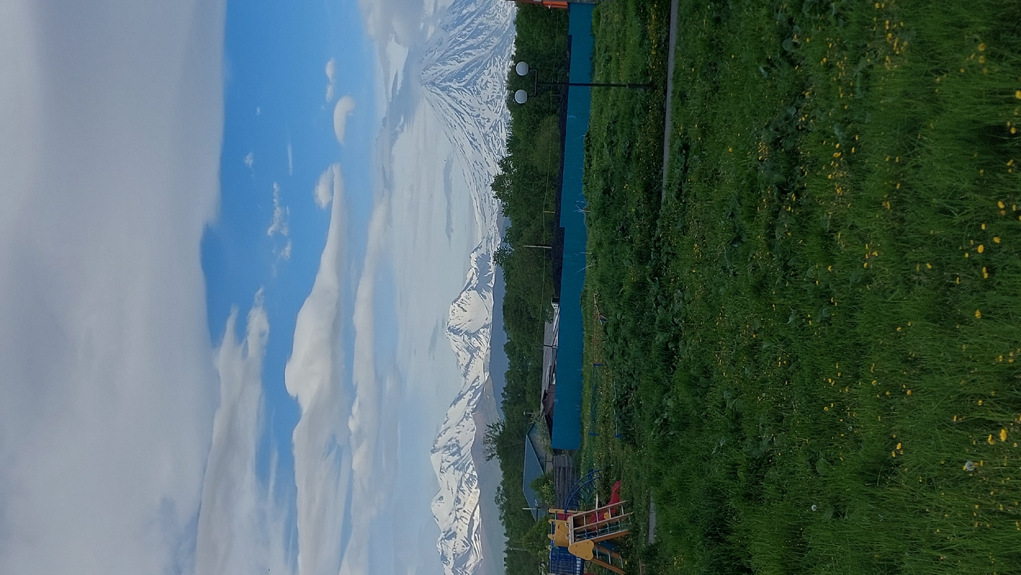The image depicts a rural landscape with a backdrop of snow-capped mountains. In the foreground, a patch of lush green grass is dotted with small yellow flowers. A children's playground structure with yellow and red elements is visible to the left. Behind the playground, a turquoise fence runs horizontally, separating the grassy area from a line of dense green trees. A few houses with dark roofs are partially visible behind the fence. In the distance, a chain of majestic, snow-covered mountains stretches across the horizon under a partly cloudy sky with patches of blue. A modern street lamp with two white globes stands near the fence. The scene appears to be in or near Зелёный, Russia, given the mountainous terrain and vegetation.