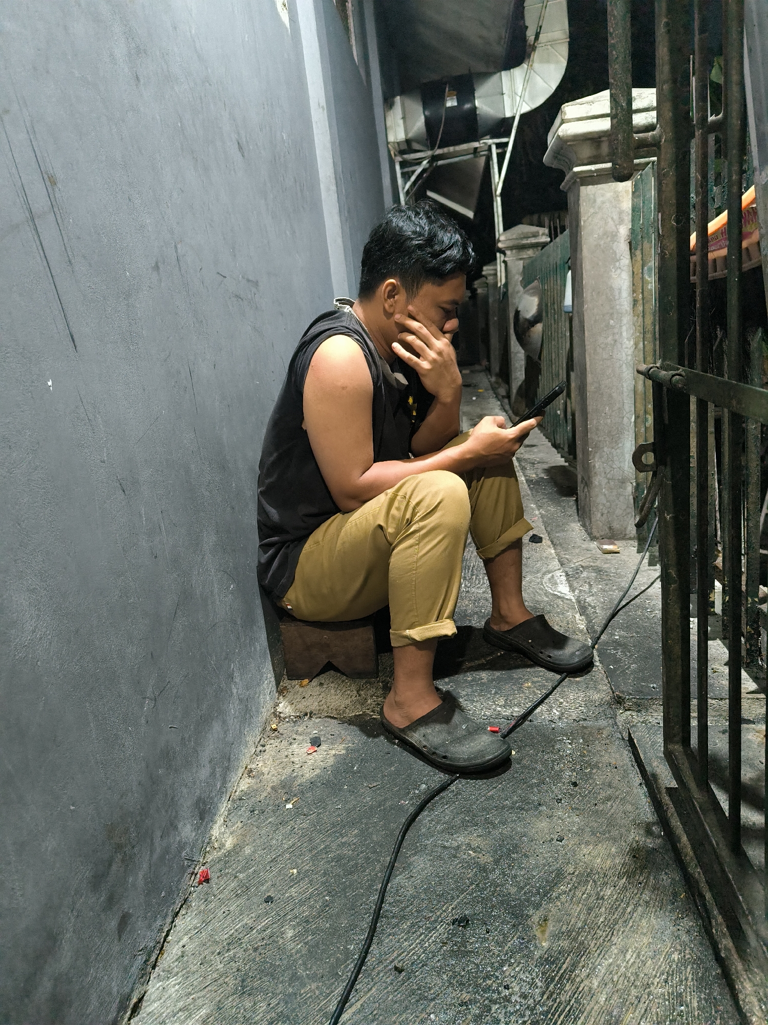 A young man is sitting on a wooden block next to a gray wall, holding a smartphone in his right hand and resting his face on his left hand. He is wearing a sleeveless black shirt and tan pants, with dark gray clogs on his feet. The ground is concrete with some debris. To the right of the man is a metal gate and a concrete pillar. The background shows an alleyway with various structures and equipment.