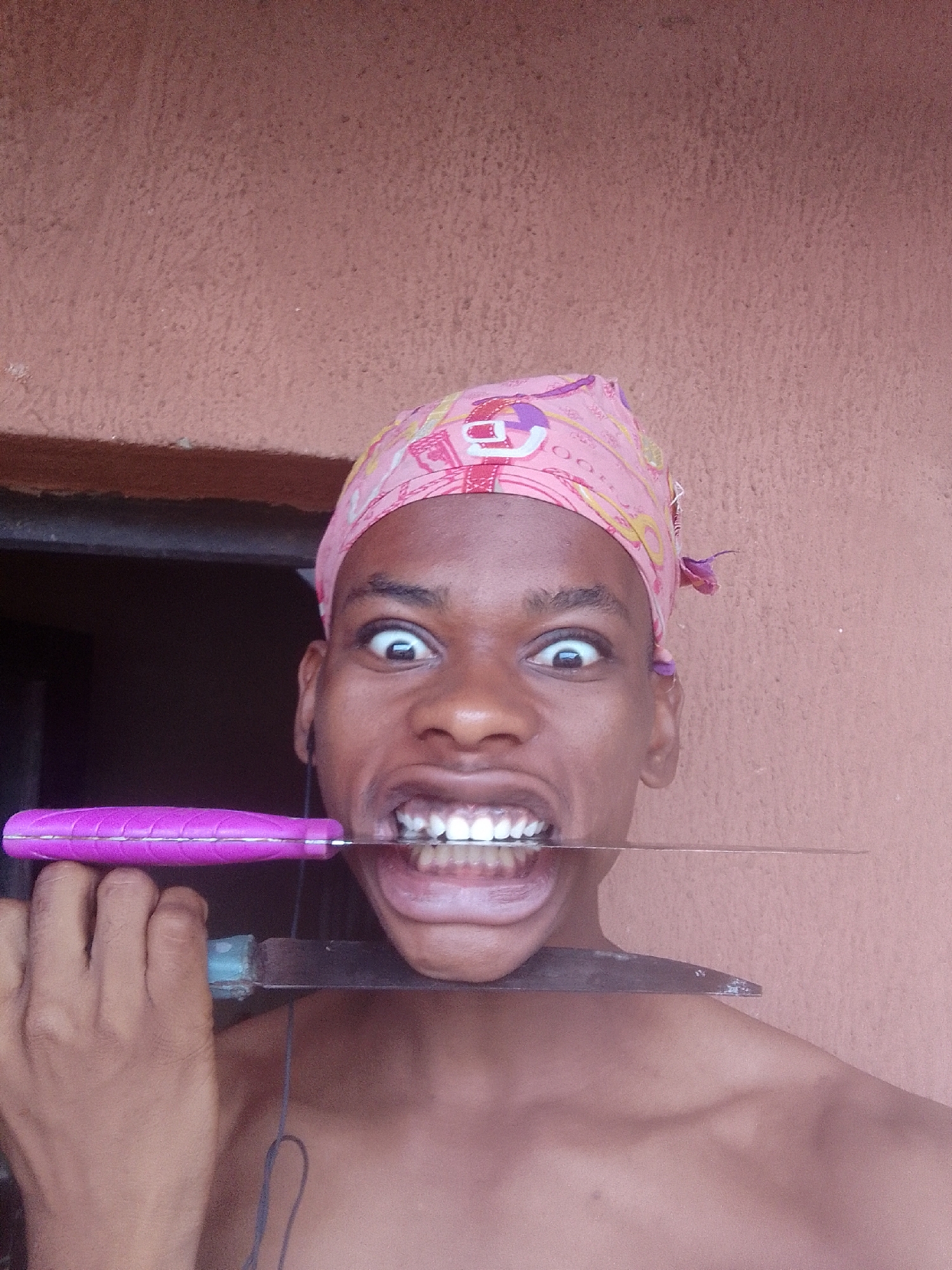 A man with bare shoulders, his head wrapped in a pink bandana, holds a large knife between his teeth, the handle gripped in his left hand. He stares directly at the camera with wide eyes. He appears to be standing near a doorframe, against a textured, reddish-brown wall. The location is Unknown, Unknown.