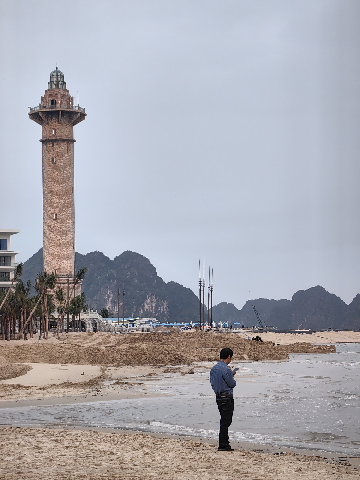 The image shows a beach scene in Bai Chay Ward, Vietnam, on an overcast day. In the foreground, a man stands at the edge of the water, looking at a mobile phone. He is wearing a blue shirt and dark pants. Behind him is a beach with piles of sand. In the midground, there are palm trees, a low white wall with arches, blue umbrellas, and three tall, thin, dark structures. A construction crane is visible in the distance. In the background, there are mountains. To the left, a tall brick lighthouse-like structure stands near a modern building. The overall environment appears to be a beach undergoing some sort of construction or renovation.