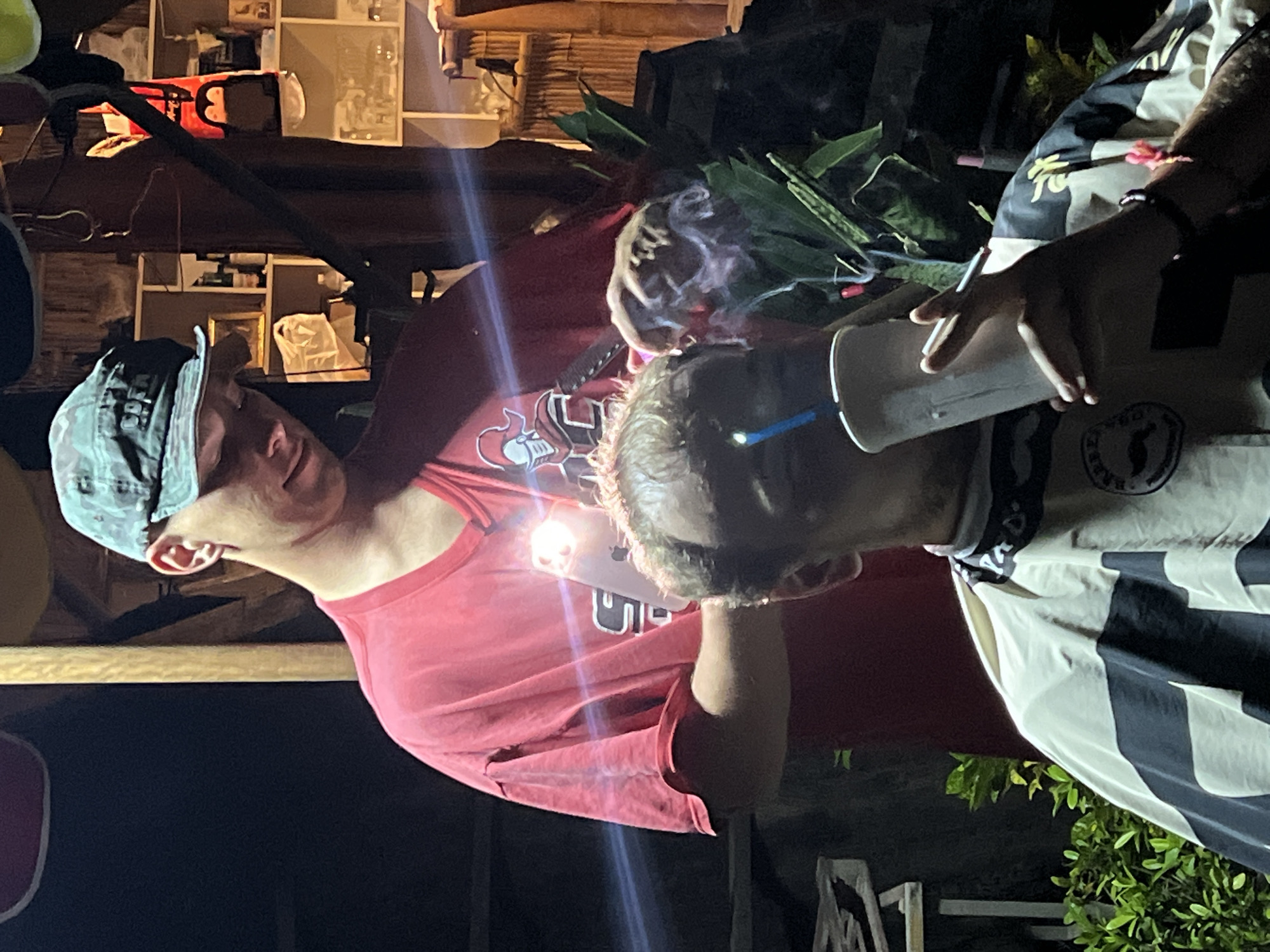 At night in an unidentified location, two individuals are positioned near vegetation. One person, wearing a red shirt and a cap, holds a phone with its light illuminated. The other individual holds a disposable cup. Smoke is visible in the scene.