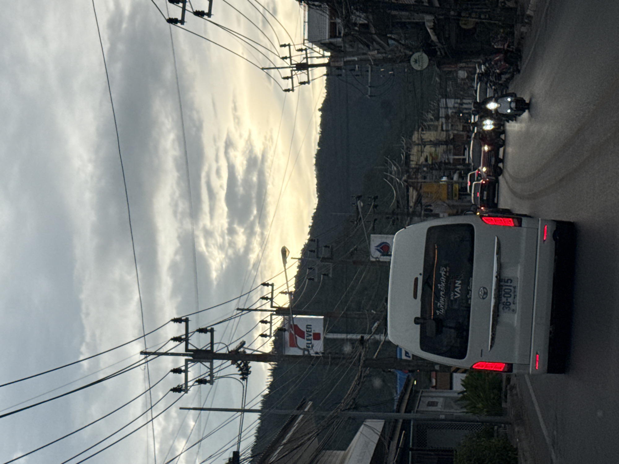 The image shows a street scene in the city of Ko Samui District, Thailand, at dusk. Power lines crisscross overhead, and telephone poles with insulators are visible. On the right side of the street, there are buildings with shops and signs, and on the left, there are more buildings and foliage. A white van is in the foreground, facing away from the camera, with its taillights illuminated. Several cars and motorcycles are on the road, with some motorcycles having their headlights on. The sky is cloudy, with patches of sunlight breaking through, suggesting the sun is low on the horizon. A "7 ELEVEN" sign is clearly visible on a pole. The overall impression is of a busy street in a Southeast Asian city during twilight.