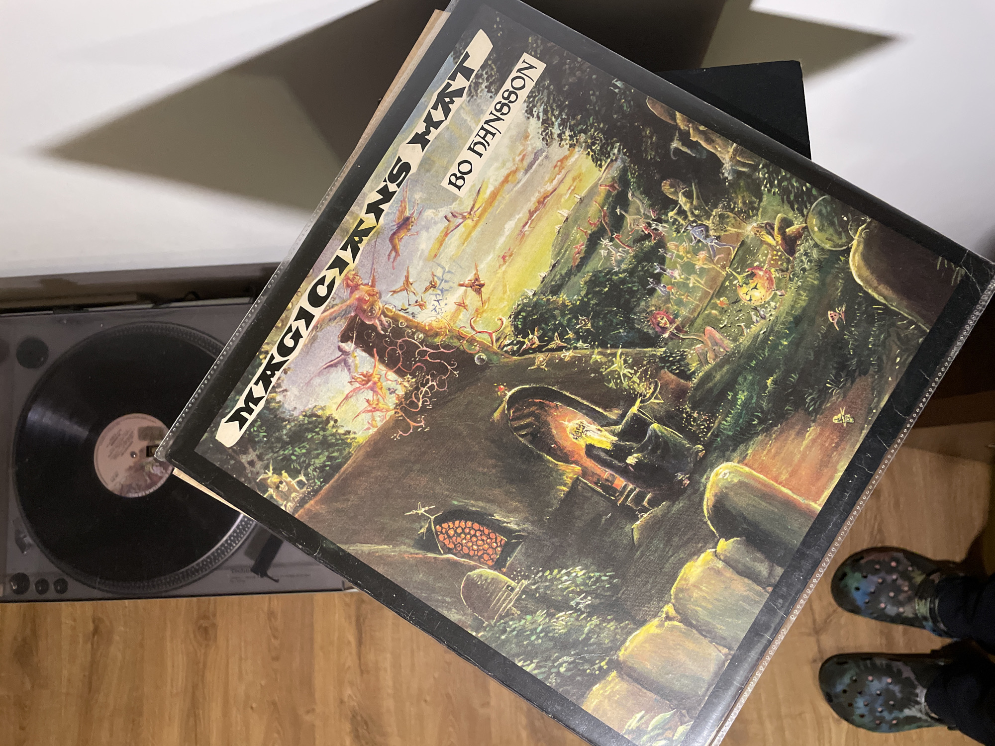 The image depicts an indoor scene in Yekaterinburg, Russia, captured from an overhead, slightly angled perspective. The primary subject is a vinyl record album titled "MAGICIAN'S HAT" by "BO HANSSON," presented within a clear plastic protective sleeve. The album cover features a detailed, fantastical landscape illustration with a dark, arch-windowed structure on the left, numerous small, whimsical figures, and a sky transitioning from dark to a sunset-like yellow and orange.
In the upper-left background, a dark gray turntable is partially visible with a black vinyl record on its platter, exhibiting a white center label. Below the album cover, at the bottom of the frame, a pair of human feet wearing dark, iridescent Crocs-style clogs are visible, suggesting the presence of an individual. The feet are positioned over light-colored wooden floorboards. A plain white wall is visible in the upper right and right side of the frame, against which the album cover appears to be leaning on a dark, unidentifiable object, casting a distinct triangular shadow on the wall. The scene is illuminated by an overhead light source, creating pronounced shadows.