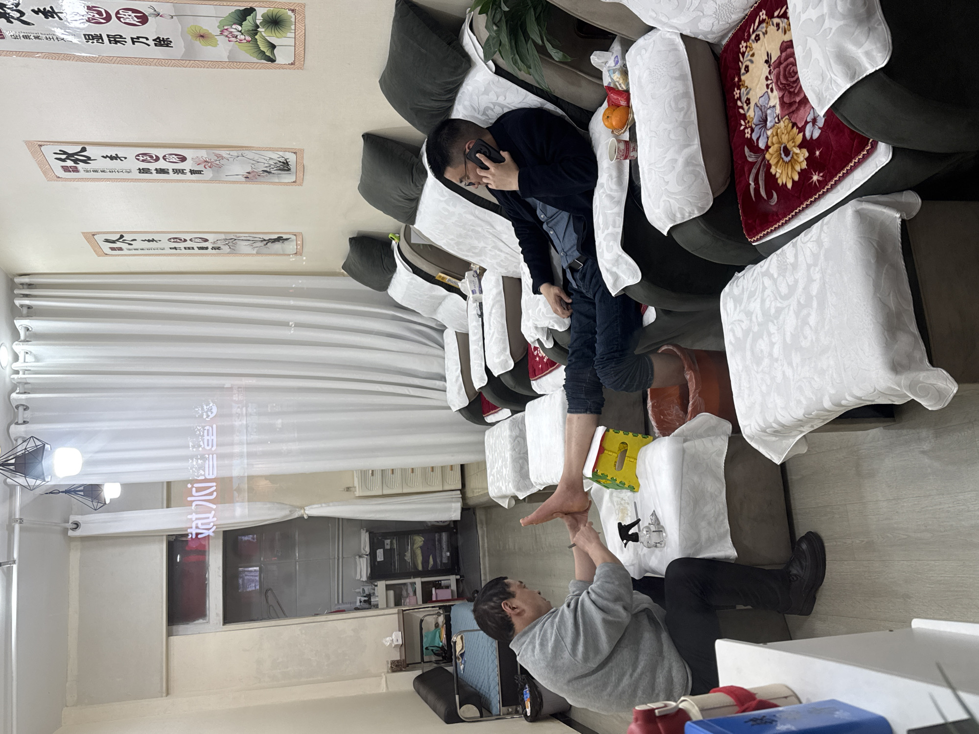 The image shows a man receiving a foot massage while talking on a phone. The setting appears to be an establishment offering massage services, possibly in Qianjiang, China, based on the traditional Chinese artwork on the walls. The man receiving the massage is seated on an upholstered lounge chair with decorative cushions and covers. The massage therapist is attending to his foot, which rests on a padded footrest. The room is divided by a sheer curtain, with a window and a kitchenette visible in the background. The atmosphere is likely meant to be relaxing and therapeutic.