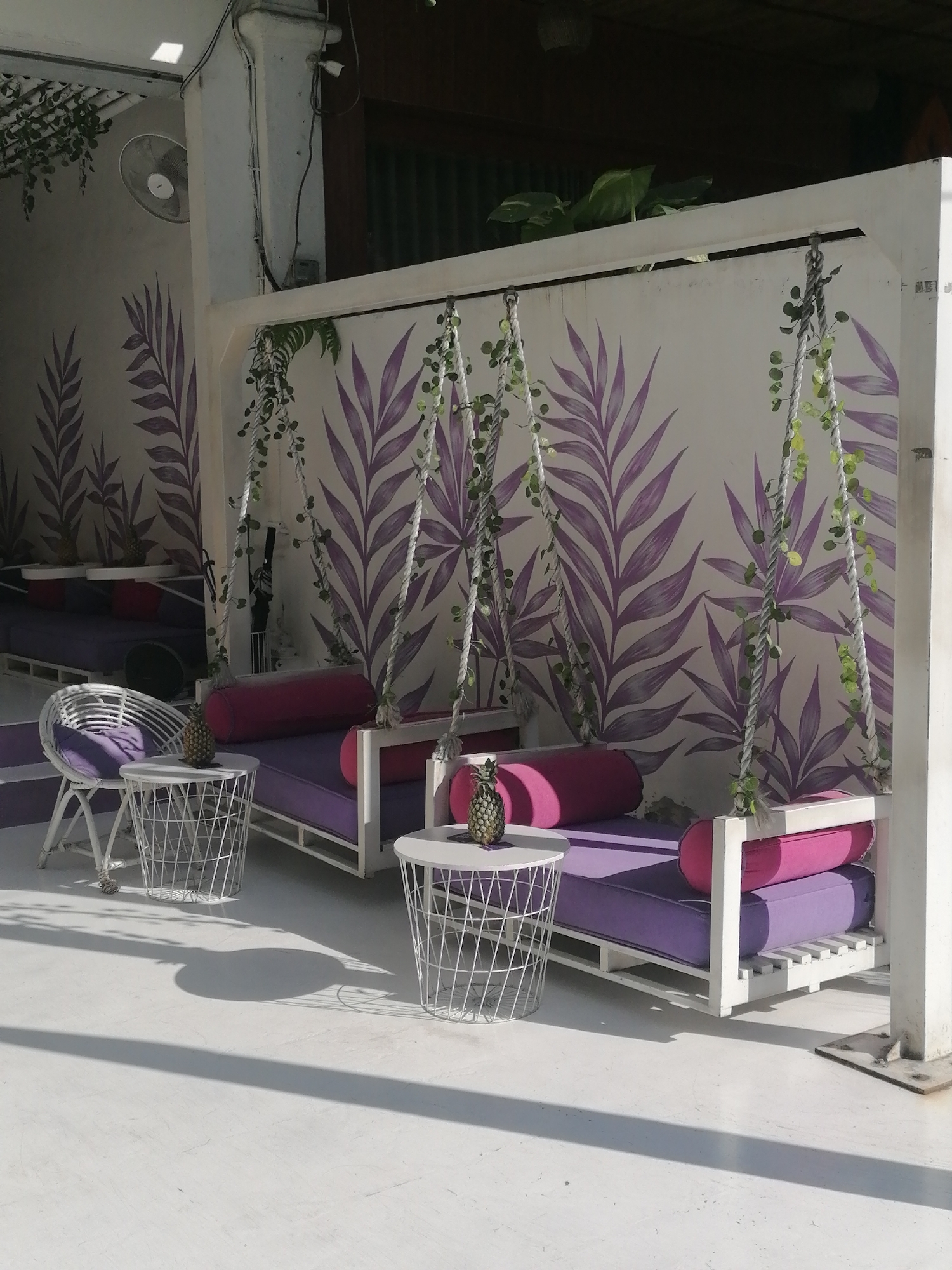 An outdoor seating area in Ubud District, Indonesia, features two white wooden swing beds with purple cushions and pink bolsters. The beds are adorned with artificial green vines. White wire mesh side tables, one with a pineapple centerpiece, are placed next to the beds. A white wicker chair with a purple cushion sits to the left, also beside a side table. The background is a white wall decorated with purple palm leaf motifs. The floor is a white, smooth surface, casting long shadows from the seating structures, suggesting it is daytime. A ceiling fan is visible on the upper left wall.