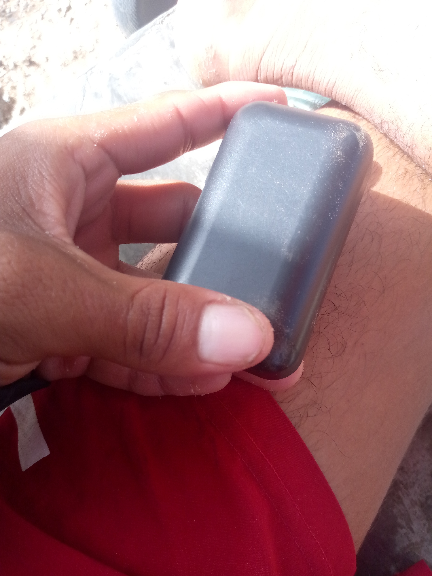 A close-up shot shows a hand holding a black rectangular object, possibly a GPS tracker or a small electronic device. The object is resting on a person's leg, which is covered in short, dark hairs. The person is wearing a red garment, likely shorts or pants. The background is blurred, but appears to be a sandy or rocky surface, suggesting an outdoor setting. The lighting is bright, with harsh shadows indicating direct sunlight. The image appears to have been taken in an outdoor urban environment.