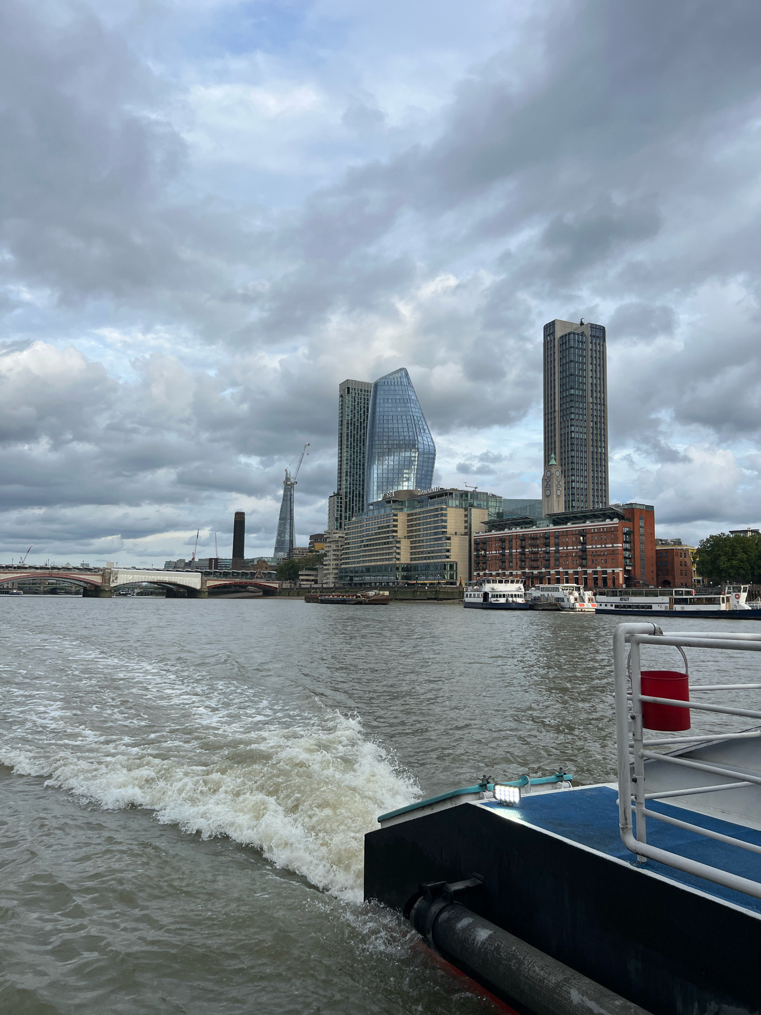 The image captures a view from a moving boat on the River Thames in London, United Kingdom. The foreground shows the wake of the boat and a portion of its deck, including a blue-painted surface, white railings, and a red bucket. In the mid-ground, the river is visible with several moored boats and ferries, including a white ferry with blue trim. Across the river, the London skyline is dominated by various buildings. Prominent structures include the distinctive shard-like building known as "The Shard," a tall, dark cylindrical building, and several modern glass skyscrapers with angular designs. A multi-span bridge with red and white accents crosses the river in the distance. The sky is overcast with heavy, grey clouds. The overall scene depicts a cloudy day in a bustling urban environment, viewed from the water.