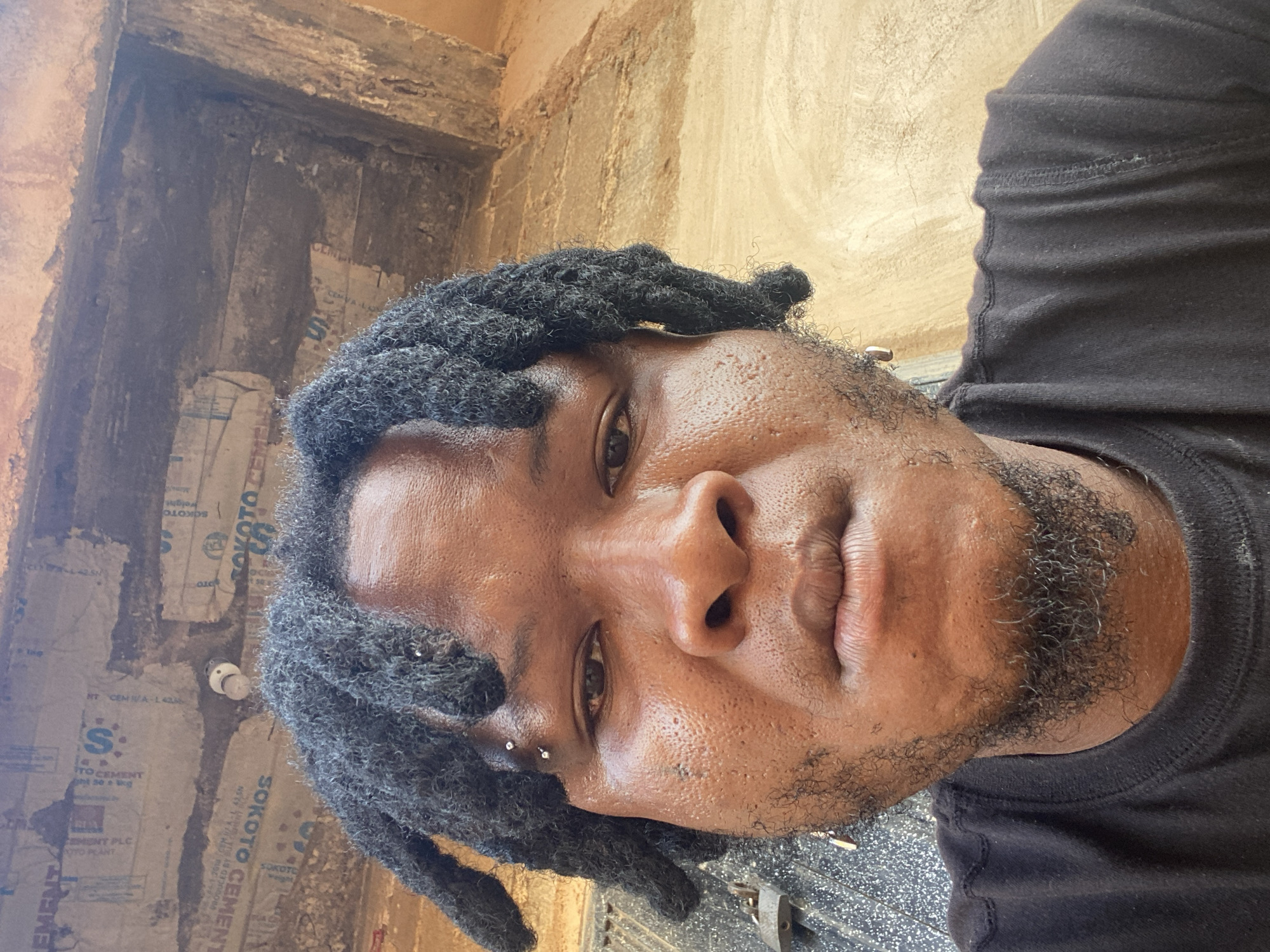 A close-up, selfie-style portrait of a Black male with dreadlocks and facial hair is shown. He is wearing a black t-shirt and has piercings on his left eyebrow and in his left ear. The background appears to be an outdoor setting with a textured wall, possibly made of concrete or stucco, and some visible scaffolding or building materials overhead. The lighting suggests it is daytime. The specific location within Gura Top, Nigeria, is not discernible from the image alone.