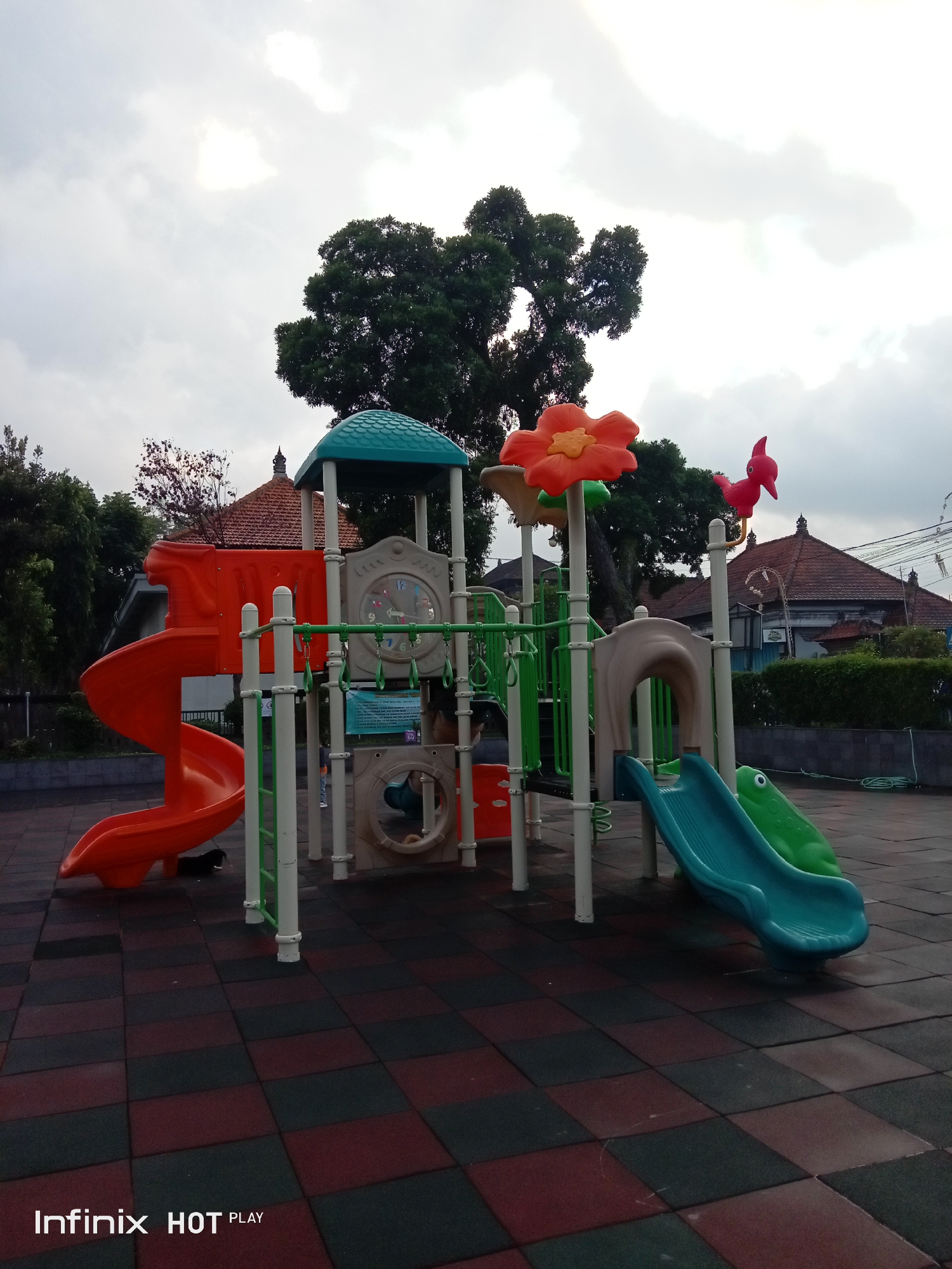 In Gianyar, Indonesia, a vibrant children's playground stands on a patterned rubberized ground. The playground features a multi-level structure with orange and green slides, a large red flower decoration, and a red bird figurine. White and green support poles frame the structure. Behind the playground, traditional Indonesian houses with terracotta roofs are visible, along with lush green trees. The sky is overcast, suggesting a cloudy day. A portion of a street with overhead wires is visible in the background. A black dog is present in the lower left portion of the image, near the orange slide.