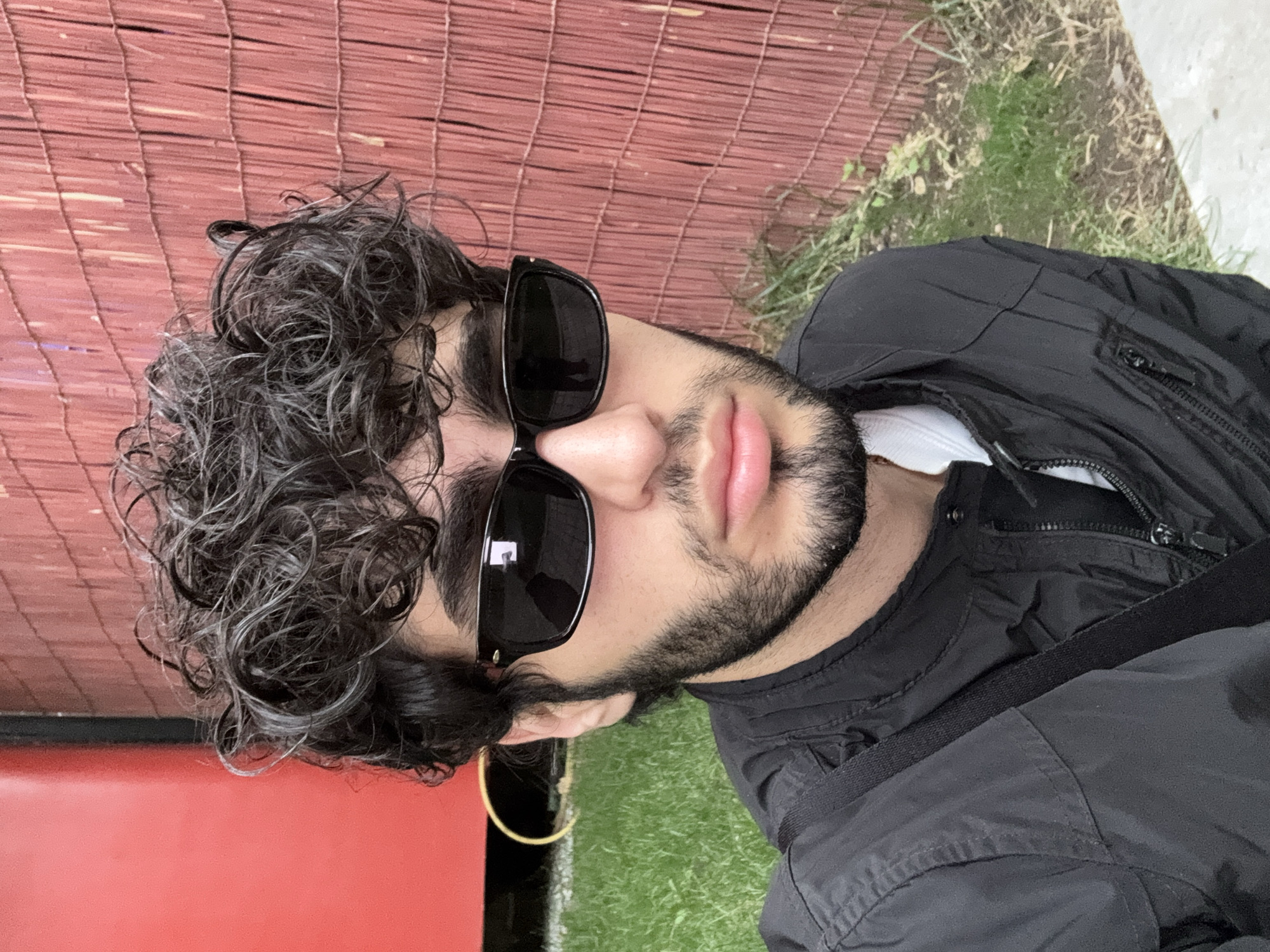 The image shows a young man with curly dark hair and a beard, wearing black sunglasses and a black jacket. He is looking towards the camera, with a slight smile on his face. The background consists of a red bamboo fence and green grass, suggesting an outdoor setting. The lighting indicates it is daytime. The man appears relaxed and casual. There is no visible text in the image. The provided location context of Yerevan, Armenia, suggests the image was taken in that city.