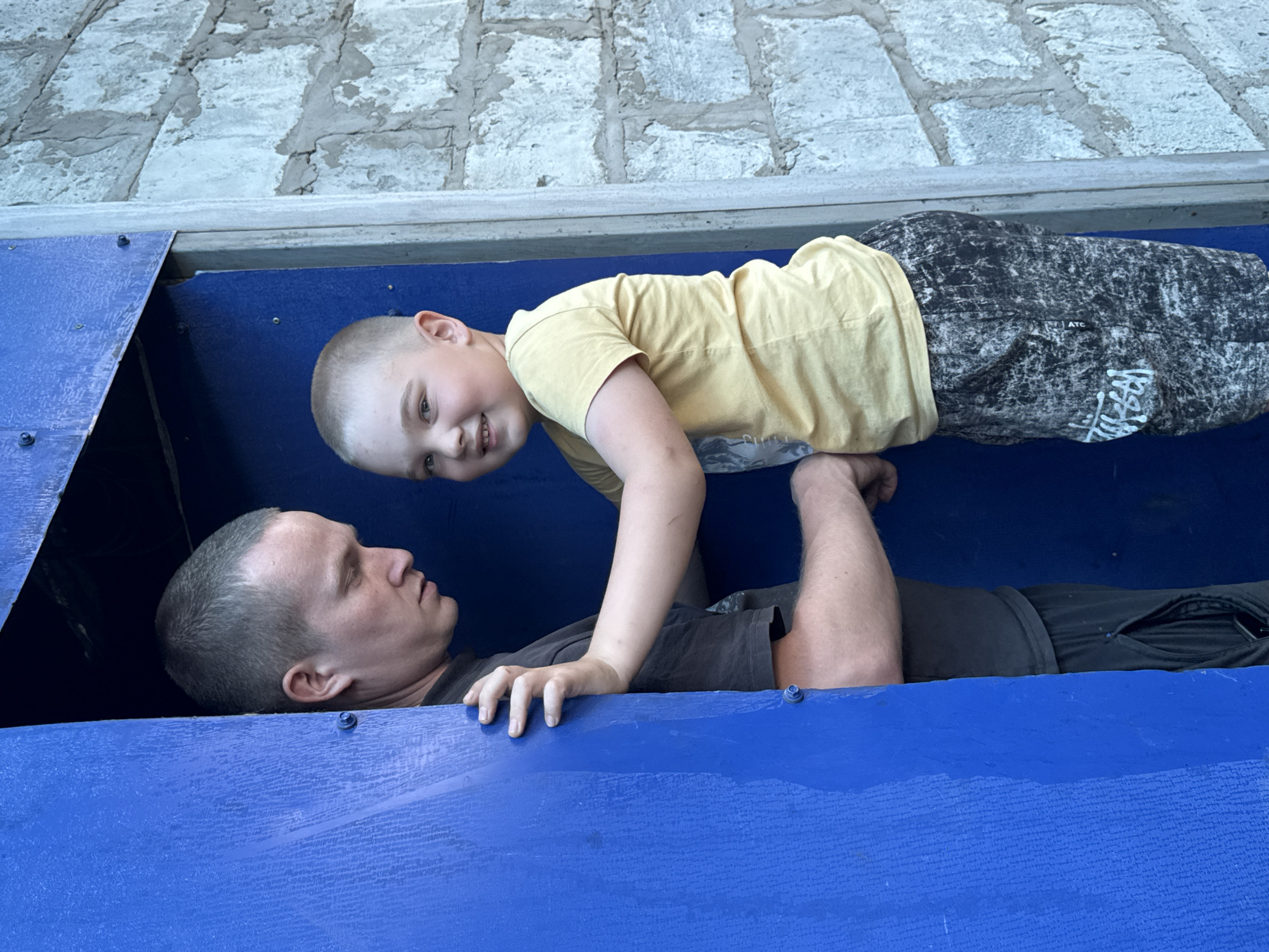 A father and son are lying in a blue box. The father is on his back, looking up at his son. The son is hovering above his father, supported by his hands and feet on the father's arms and legs. The son is wearing a yellow shirt and patterned pants and is smiling. The father is wearing a gray shirt and black pants. The box is located outside near a concrete structure. Based on geolocation information, this image was taken in the city of Zhigalki, Russia.
