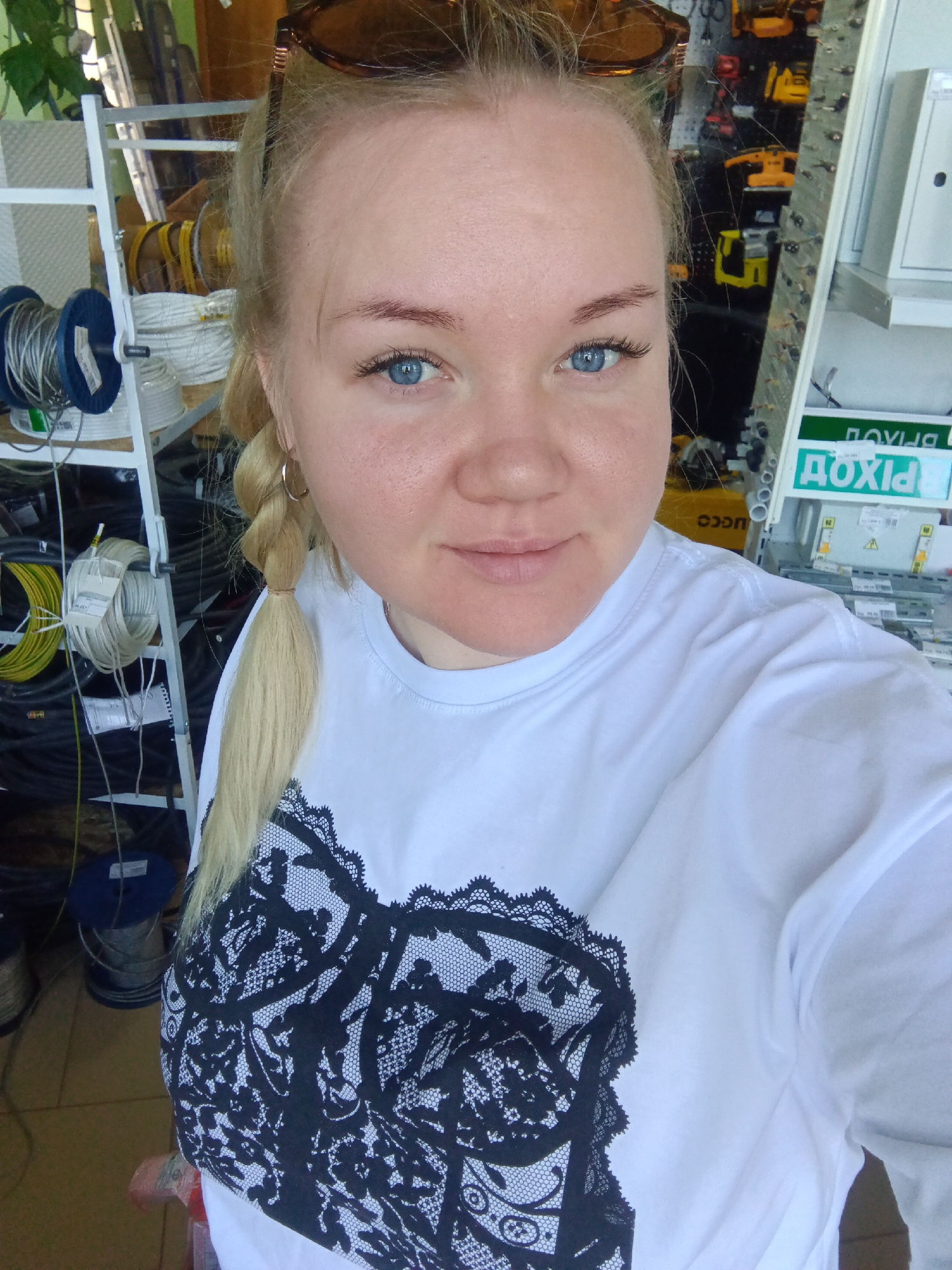 A fair-skinned woman with light blonde hair in a braid stands indoors, taking a selfie. She has blue eyes and is wearing a white t-shirt with a black lace-like design on the front. Sunglasses rest on top of her head.
Behind her are shelves stocked with spools of various wires and cables. Tools and equipment are visible in the background, along with signage in Russian ("ДОХІД"), suggesting a shop or workshop environment. The location may be Фоки, Russia, given the city request, but this is not definitively confirmed by the image alone.