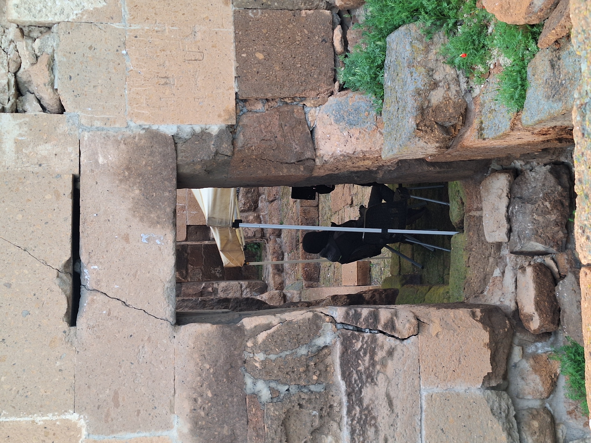 A photograph shows a view through a rectangular opening in a stone structure. The structure appears to be part of the ruins, with uneven, cracked, and weathered stone blocks forming the opening's frame. Small patches of green vegetation grow on top of some of the stones. Through the opening, a set of stone steps can be seen, with a person sitting on a chair is visible on the steps. The photograph was taken in the city of Anipemza, Armenia.