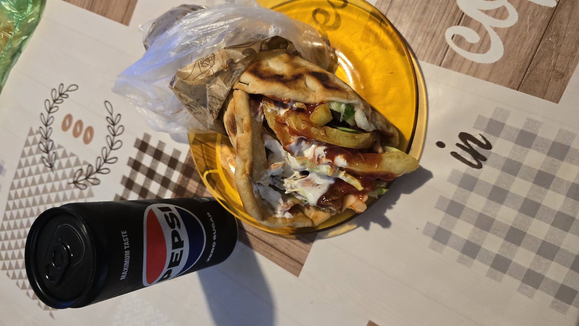 An overhead shot shows a black can of Pepsi Zero Sugar next to a yellow plate with a gyro and fries. The gyro is wrapped in pita bread and stuffed with meat, fries, salad, and sauce. The Pepsi can is black with the Pepsi logo in red, white, and blue. The plate is a bright yellow and is set on a tablecloth with a checkered pattern. In the background, there is a decorative element with leaves and circles. The word "in" is visible on the tablecloth in a dark script font. The scene appears to be set on a table.
