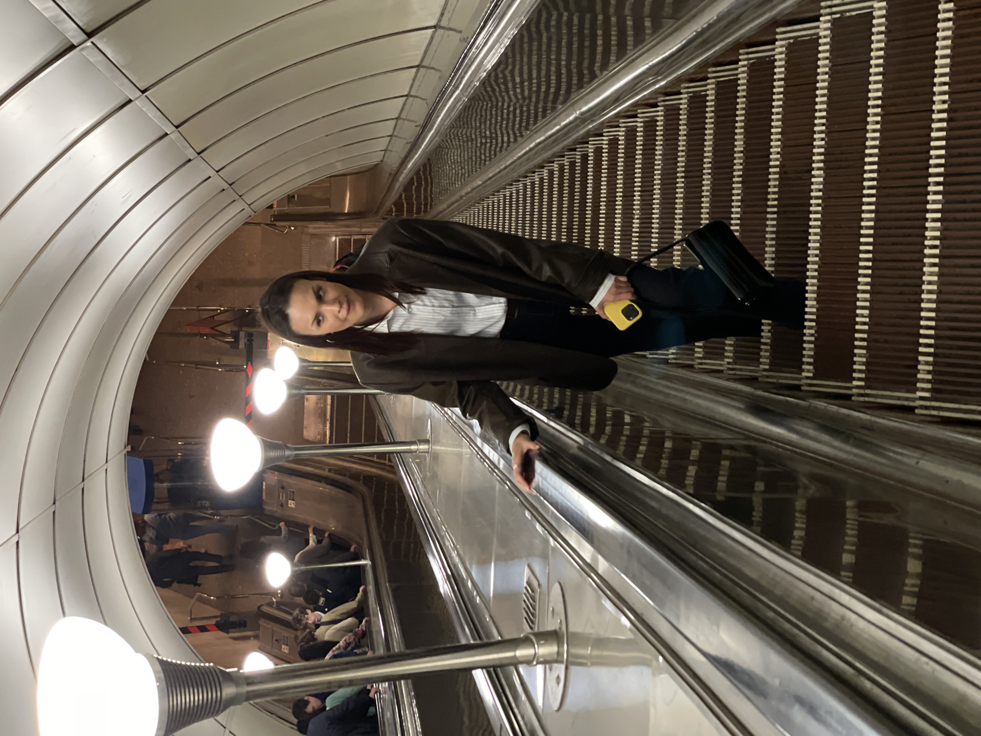 A woman stands on an escalator in an underground station. She is looking toward the camera and has a mobile phone in her hand. Other people are visible standing on the escalator behind her, headed in the same direction. The escalator ascends through a curved, white-tiled tunnel illuminated by spaced-out globe lights.