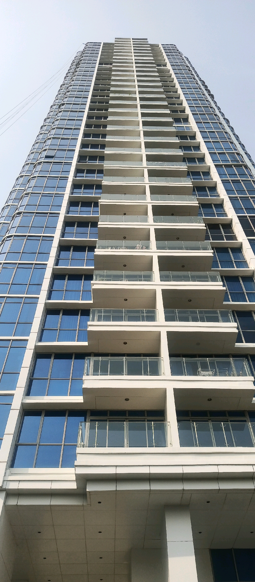This is a low-angle, full-shot photograph of a modern skyscraper in West Jakarta, Indonesia. The building is characterized by its sleek, contemporary design featuring a facade of alternating glass windows and white balconies. The glass panels are a deep blue, reflecting the sky. The balconies are staggered and appear to be a uniform width, with clear glass railings supported by thin metal posts. Some balconies are empty, while others contain minimal outdoor furniture, suggesting occupancy. The building rises vertically, dominating the frame and reaching towards a clear, pale blue sky. The perspective emphasizes the sheer height and imposing nature of the structure. The lower portion of the image reveals the base of the building with a tiled ceiling and recessed lighting.