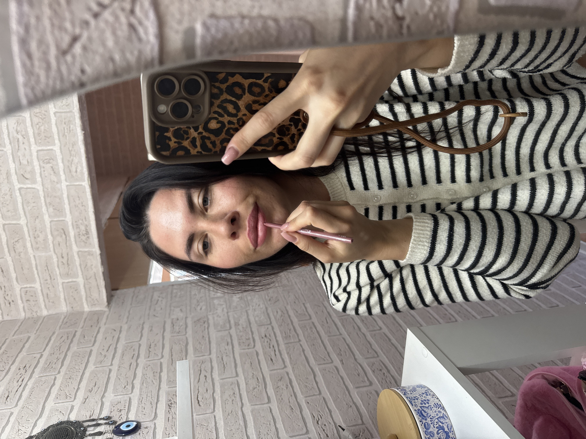A woman with dark hair and light skin is applying lipstick with a pink pencil in front of a brick-patterned wall. She is wearing a black and white striped sweater and holding a phone with a leopard-print case. The scene is indoors, with a white shelf on the left side of the image. There is no indication of the city of Chaikovsky, Russia.