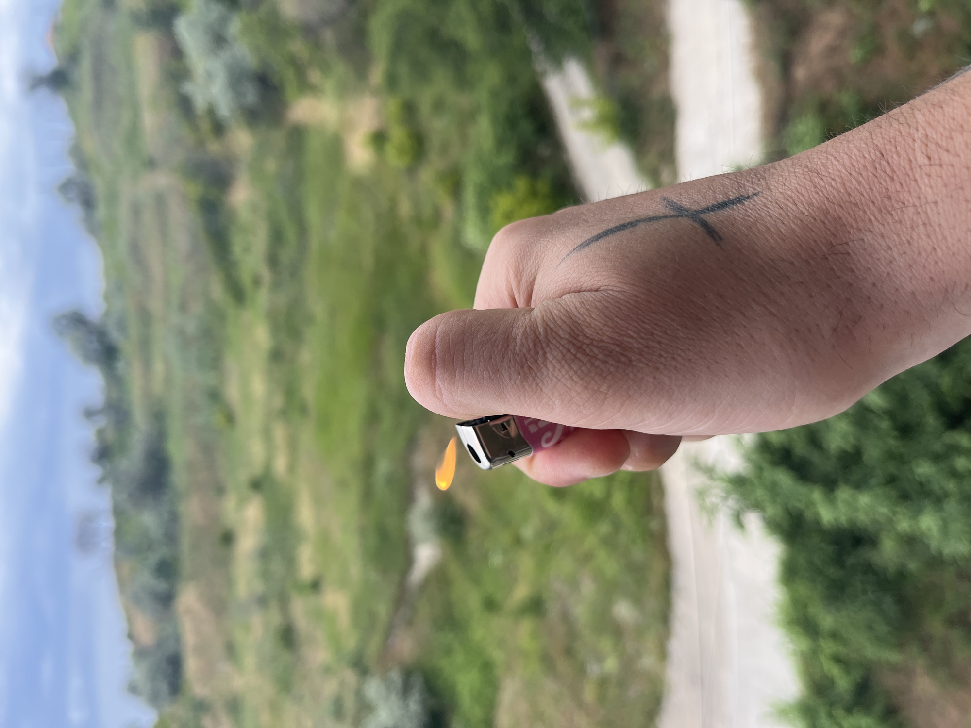 A close-up, eye-level shot shows a hand holding a pink and silver lighter with a small orange flame emerging from it. The hand has a black tattoo on its back that resembles a cross or an hourglass. The background is a blurred landscape of green hills with a cloudy sky. This image was taken in the city of Varna, Bulgaria.