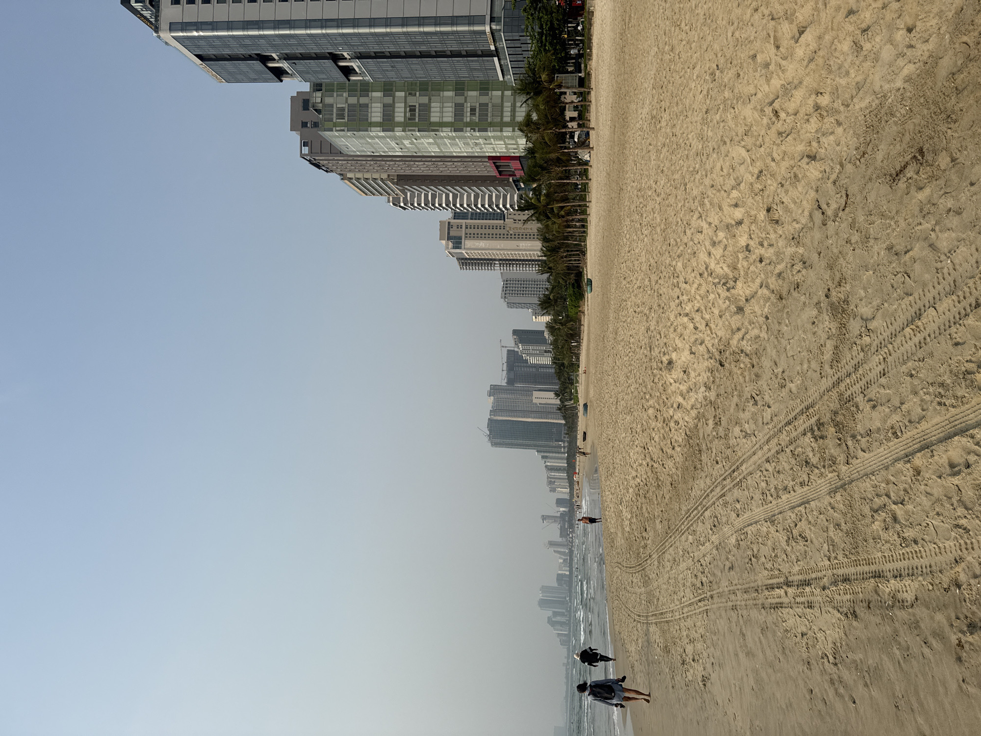 The image depicts a beach scene in Đà Nẵng, Vietnam, with a line of high-rise buildings visible along the shore. A sandy beach takes up much of the foreground, marked with tire tracks. Two individuals are walking near the water's edge, while other people can be seen further down the beach. The sky is clear and the atmosphere appears hazy.