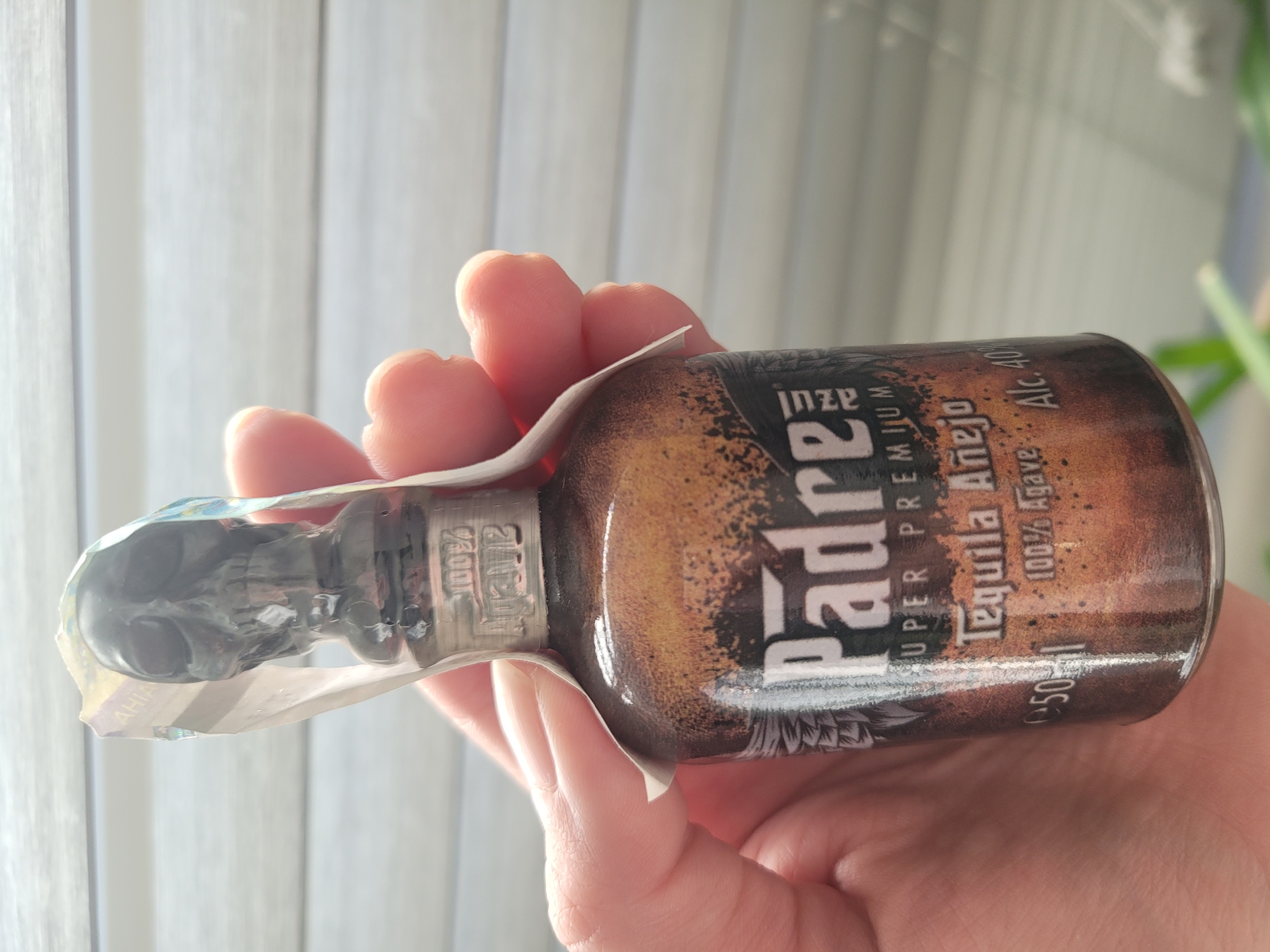 The image shows a hand holding a small bottle of "Padre" Tequila Añejo. The bottle has a skull-shaped cap. The label on the bottle is dark with brown and gold accents and reads "Padre SUPER PREMIUM Tequila Añejo 100% Agave Alc. 40%". The background appears to be a set of closed blinds. The image was taken in Kyiv, Ukraine.