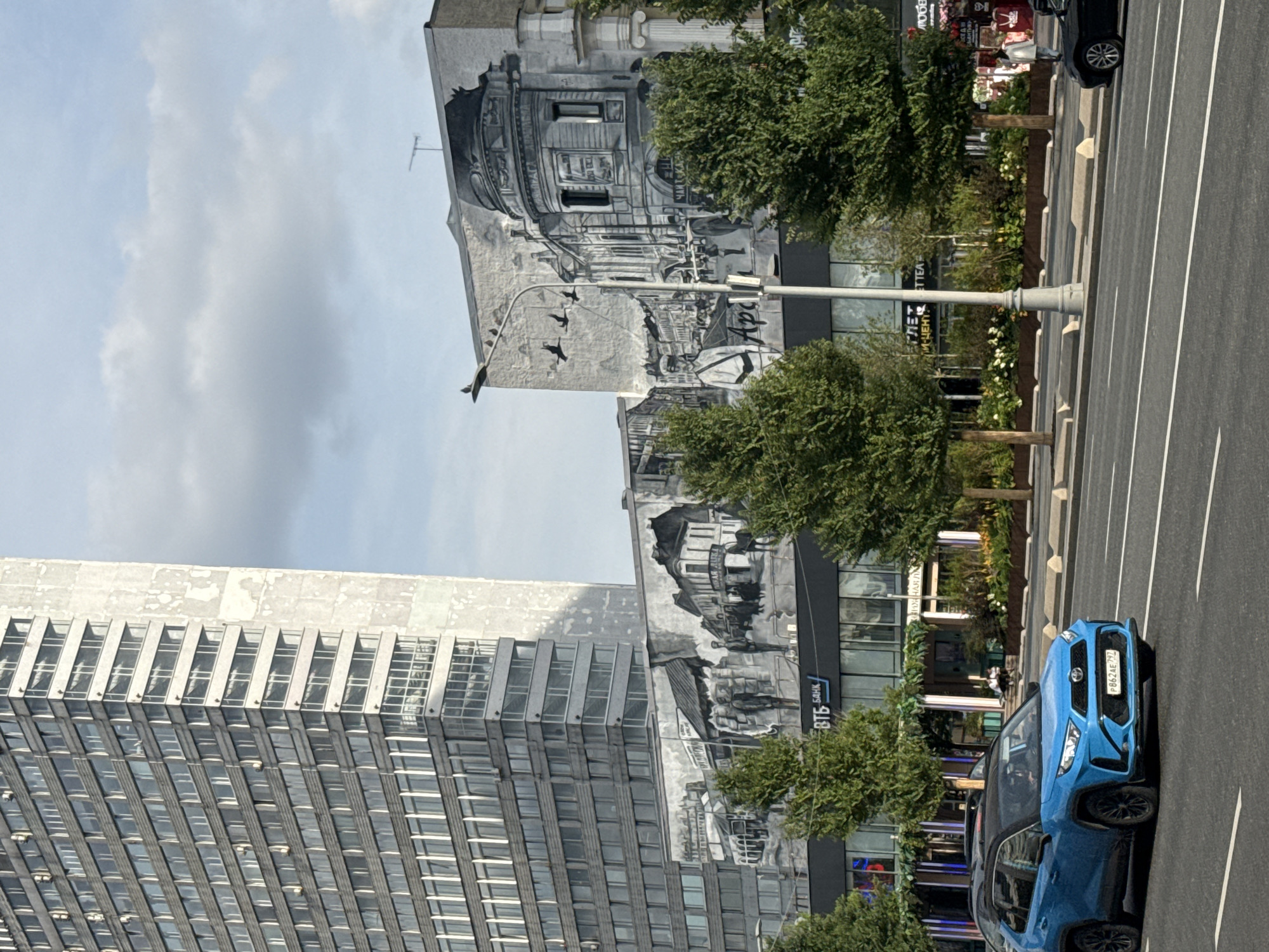In Moscow, Russia, a blue Toyota RAV4 drives on a city street. Behind the car, a large building is adorned with a black and white mural depicting historical scenes. The mural shows a street with people and horse-drawn carriages, as well as an ornate building with a dome. A tall, modern skyscraper stands to the left of the mural. The sky is partly cloudy.