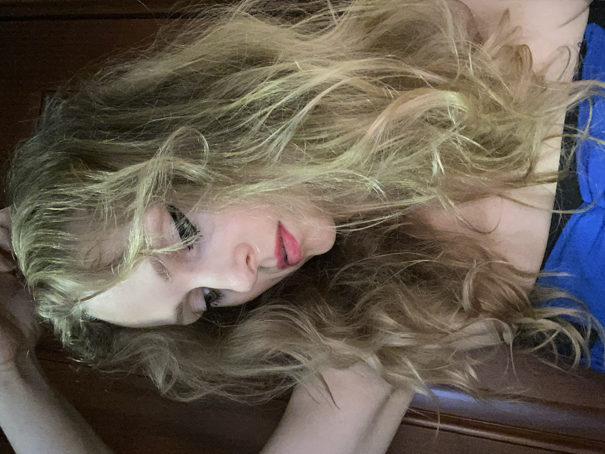 The image shows a close-up of a young woman with long, wavy blonde hair. She is wearing a blue garment. Her head is tilted down, and she is looking away from the camera. Her makeup includes winged eyeliner and pink lipstick. The background appears to be a wooden surface. There are no discernible elements that would indicate a specific location within the city of Lida, Belarus.