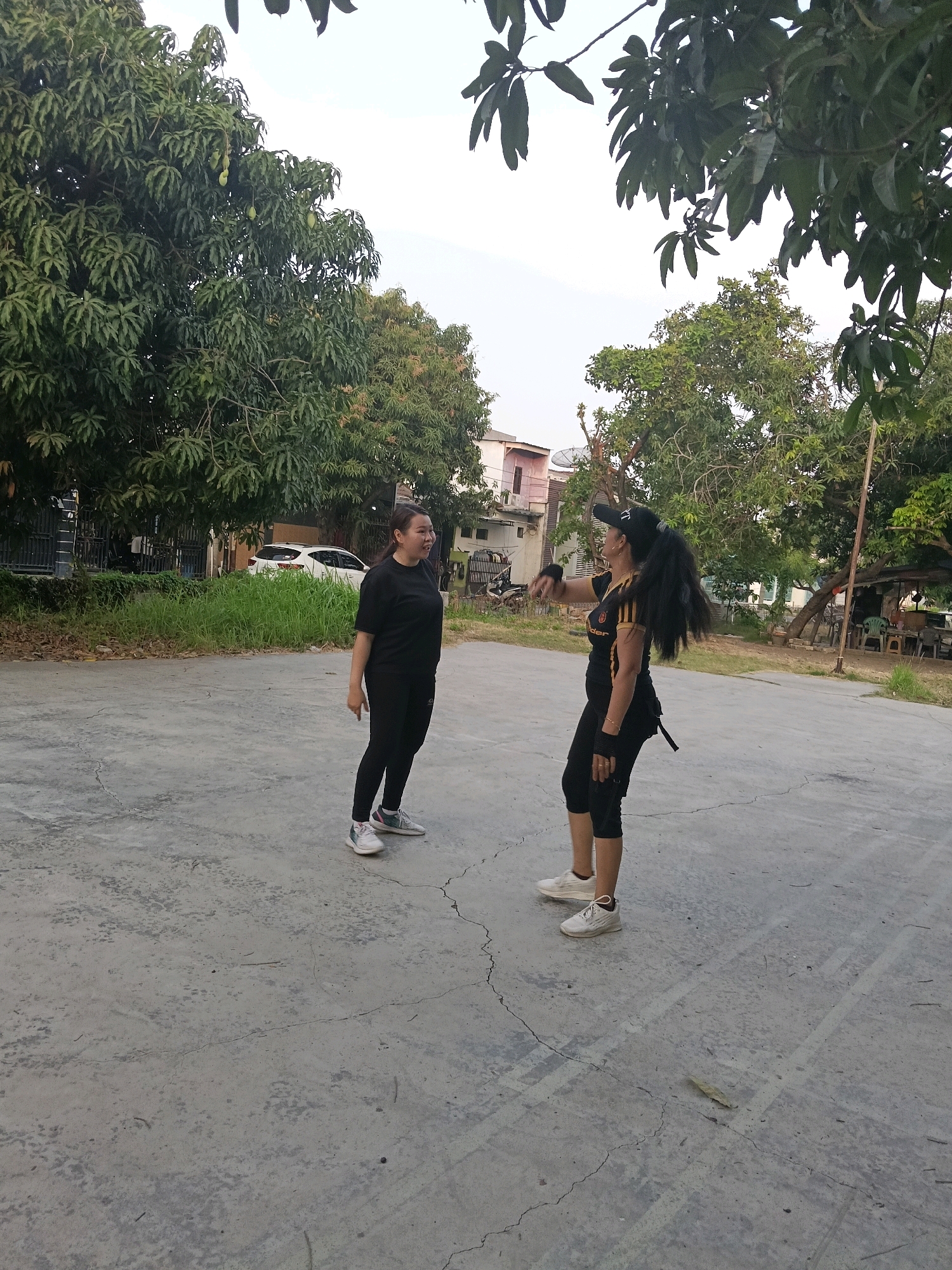 Two women are standing on a concrete court, possibly in Jatimulya, Indonesia. The woman on the left is wearing a black t-shirt and black leggings with white and grey sneakers. She is looking to her right with a slight smile. The woman on the right is wearing a black cap, a black and yellow athletic shirt, black capri leggings, and white sneakers. She is looking to her left and gesturing with her right hand. Her long dark hair is pulled back in a ponytail. In the background, there are trees, grass, a white car, a motorcycle, and buildings. The sky is overcast.
