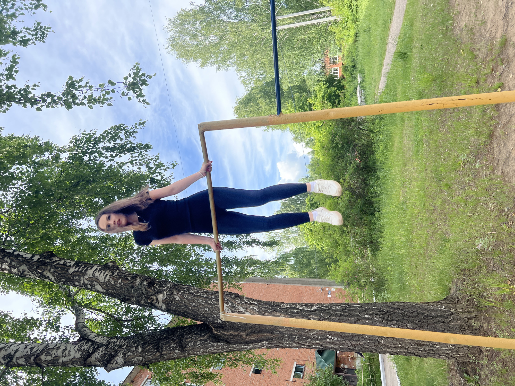 A young woman is captured mid-exercise on an outdoor horizontal bar structure, performing what appears to be a front lever or similar calisthenics move. She is positioned horizontally, parallel to the ground, holding onto the yellow-painted metal bar with both hands. Her body is straight, and her legs are extended, maintaining a strong, focused pose.
She is dressed in an athletic black t-shirt, black leggings, and white sneakers. Her long, dark hair falls over her shoulders. The setting is an outdoor recreational area, possibly a park or sports ground, characterized by lush greenery and natural light.
In the foreground and mid-ground, a large, mature tree with distinctive white and black patterned bark (birch tree) stands prominently, its branches extending towards the top of the frame. The ground beneath is a mix of green grass and patches of dirt.
The background features more trees and dense foliage, indicating a natural environment. A reddish-brown brick building with several windows is visible in the distance, partially obscured by the trees. The sky above is bright blue with scattered white clouds, suggesting a clear day, likely morning or afternoon. There is no visible text in the image. The overall impression is one of athletic effort in a verdant, open-air setting in Noviy, Russia.