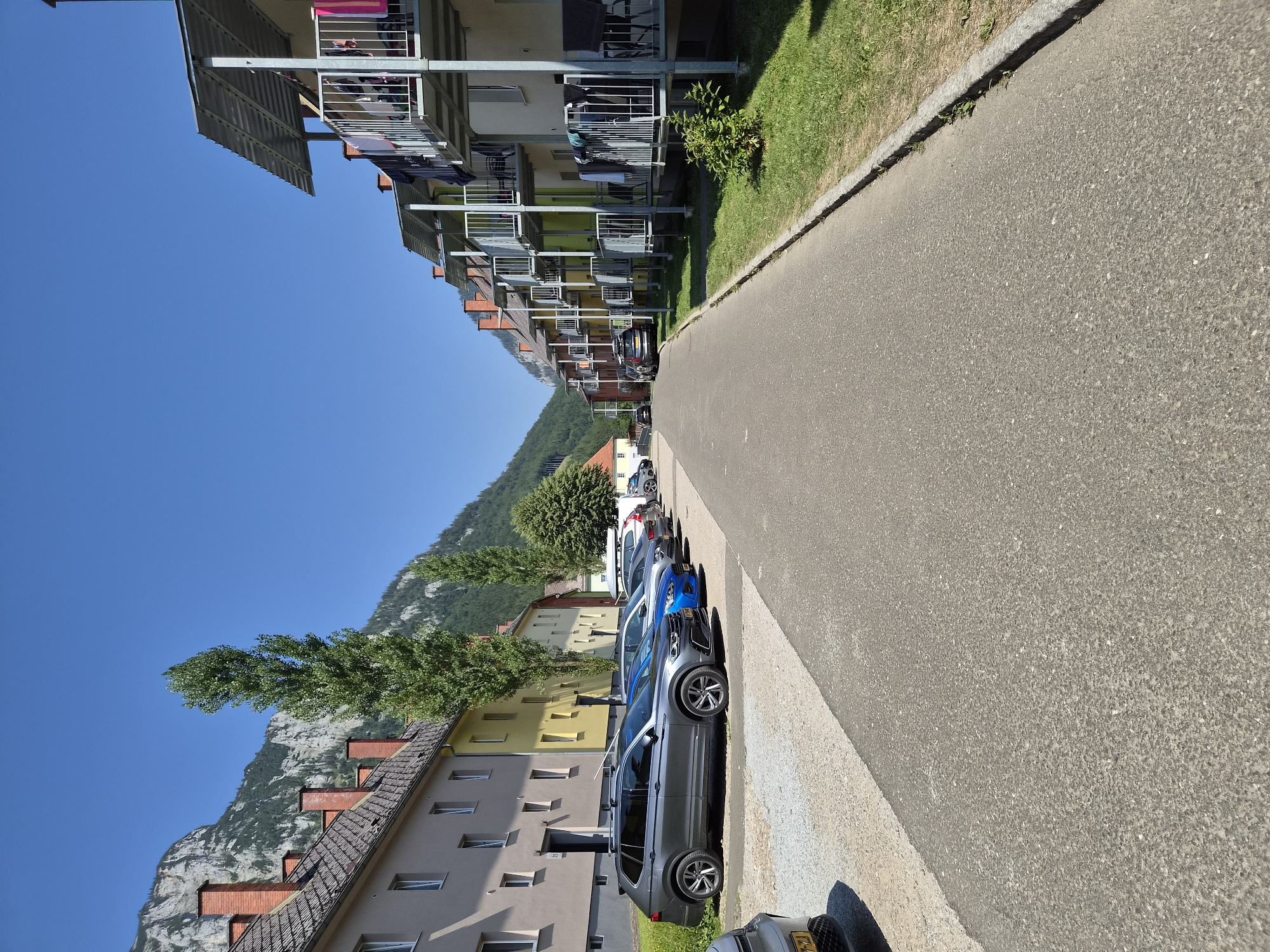 The image depicts a residential street in the city of Eisenerz, Austria, with apartment buildings on either side of a paved road. The buildings on the left are a row of apartment blocks with light-colored facades and red-tiled roofs, some with visible chimneys. A line of parked cars is situated in front of these buildings, including a dark gray SUV in the foreground. Tall, slender trees stand between the buildings and the road.
On the right side of the road, another row of apartment buildings with balconies is visible. These buildings have multi-colored facades, ranging from pale yellow to green and pink. The balconies are equipped with metal railings and some have laundry hanging out to dry. The street is lined with a patch of grass and a sidewalk curb. In the background, a steep, rocky mountain range with some vegetation is prominent against a clear blue sky. The scene appears to be a sunny day. No people are visible in the image.