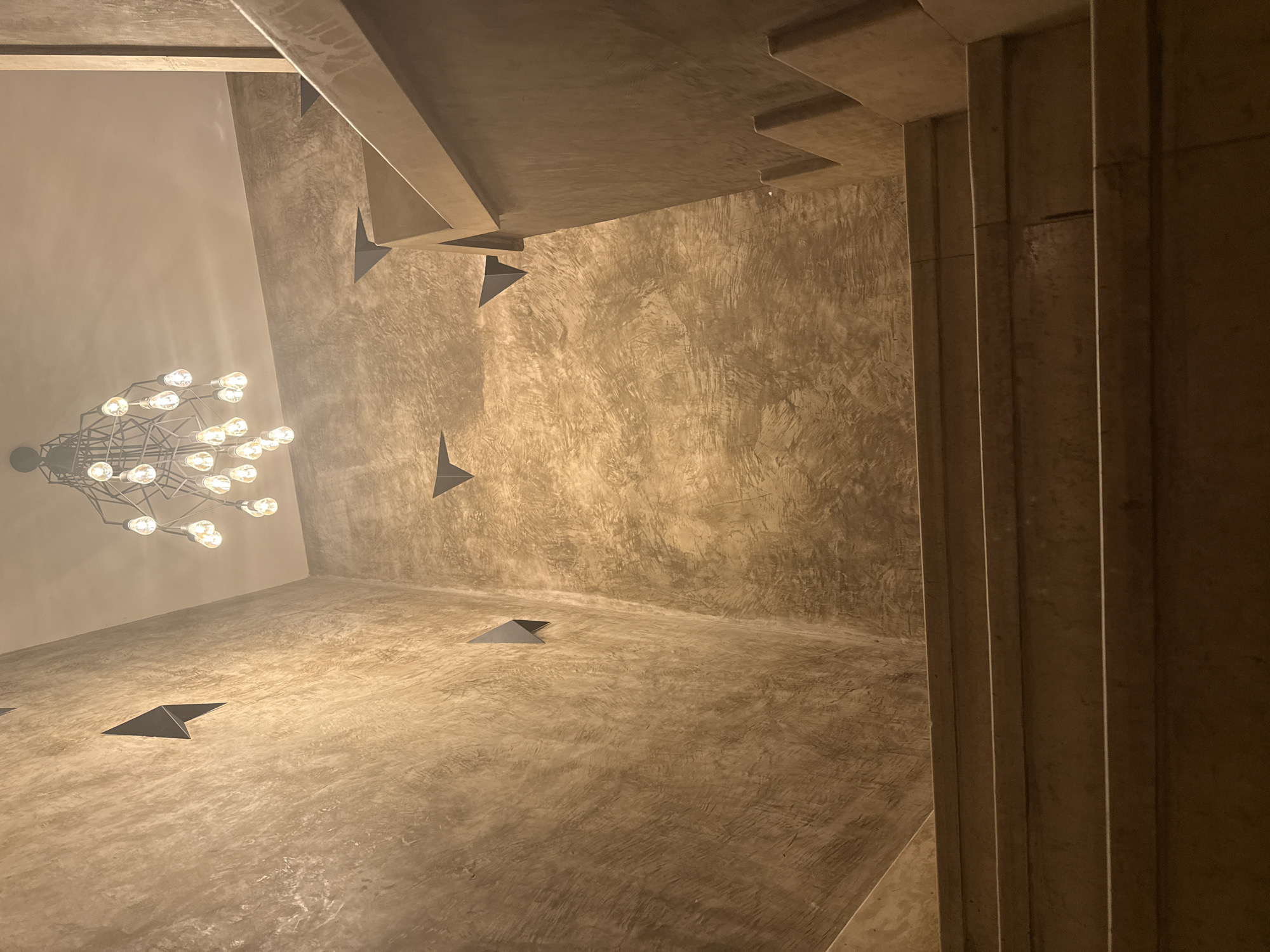 This is a low-angle, interior shot of a staircase with textured, concrete-like walls and a modern chandelier hanging from the ceiling. The staircase appears to be made of light-colored stone or concrete. Several triangular, dark grey architectural elements protrude from the walls at various heights. The chandelier is a complex geometric structure with numerous exposed light bulbs, which are illuminated. The lighting is warm and casts shadows. The scene appears to be set in a contemporary building with a focus on industrial or minimalist design. There is no indication of Varna, Bulgaria, in the image itself.