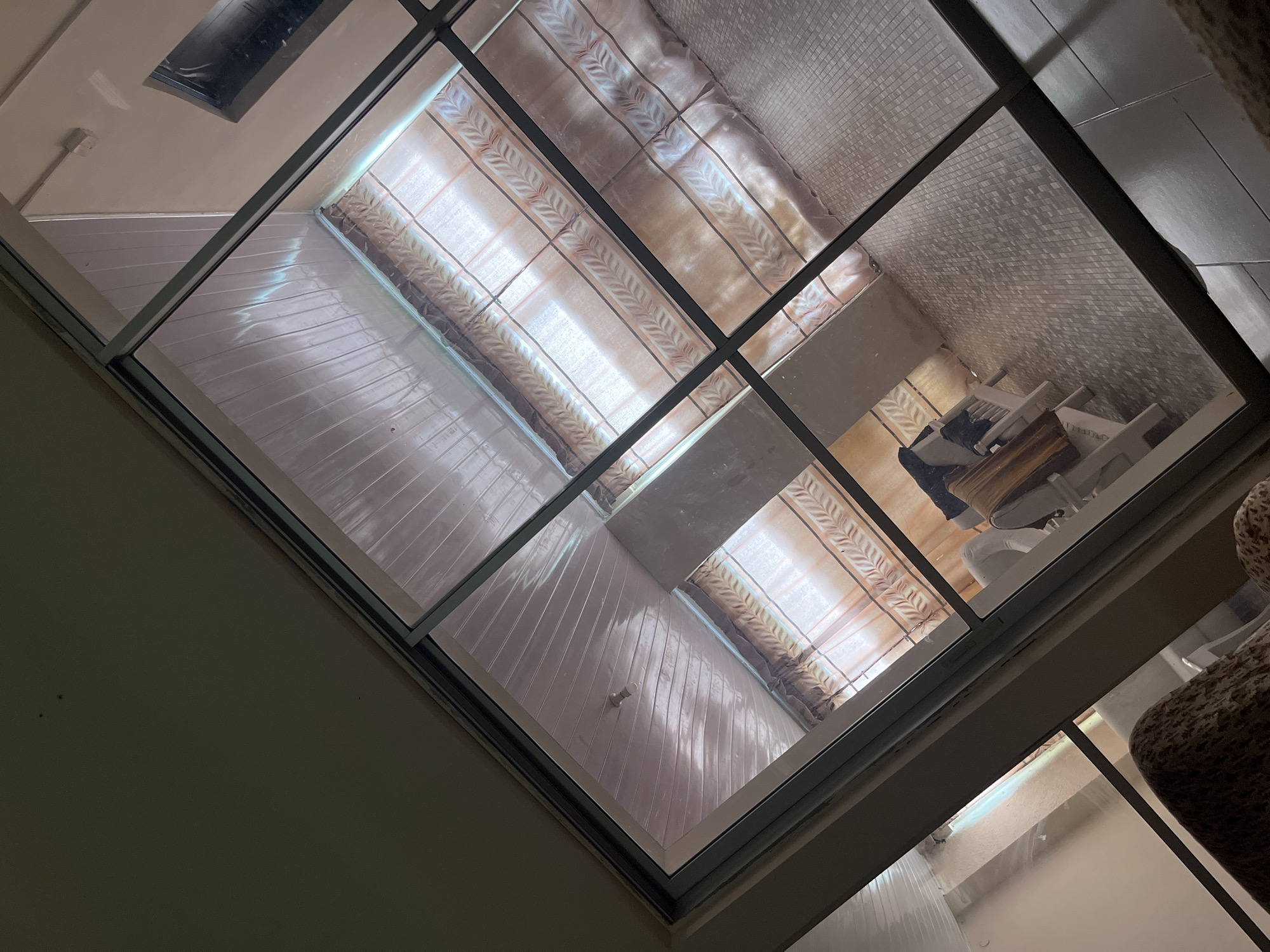 The image is a high-angle shot looking down through a glass ceiling or skylight into a room. The room appears to be an enclosed patio or sunroom with a white, wood-paneled ceiling. There are beige curtains with a subtle patterned design hanging behind the glass. In the room, there is a small wooden table with two white chairs. A dark piece of fabric or clothing is draped over one of the chairs. The floor is tiled with small, square mosaic tiles in a gray and beige pattern. The overall lighting in the room is dim. The scene is viewed from what appears to be an interior space, looking out and down. There are no specific details that indicate a location in Zawan, Nigeria.