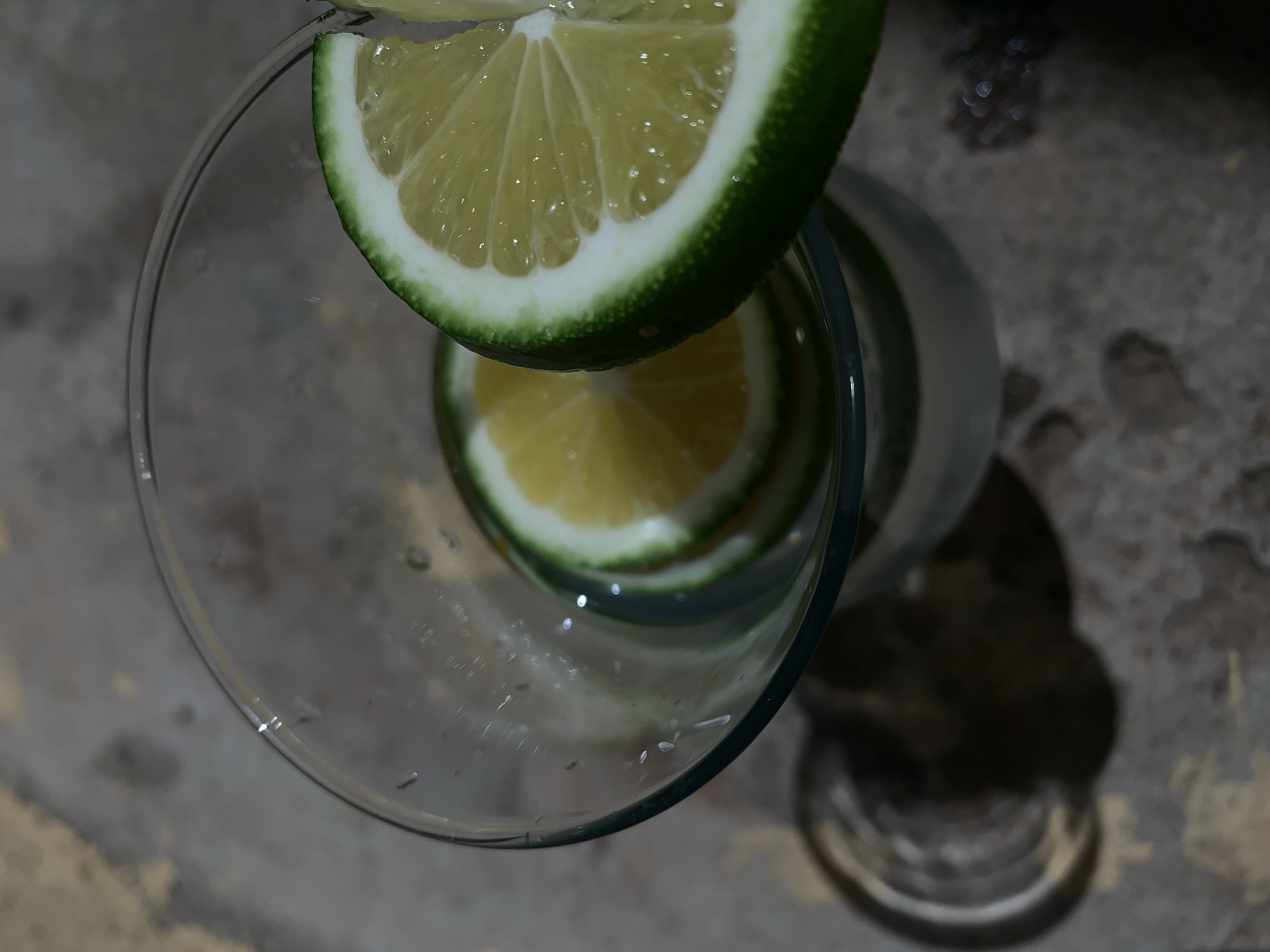 A high-angle close-up shot shows a clear glass with two lime slices. One slice sits on the rim of the glass, while the other is inside. The environment is dimly lit and appears to be a rough surface, casting shadows. The location, Rafin Par, Nigeria, cannot be verified from the image. No people, actions, or interactions are visible.