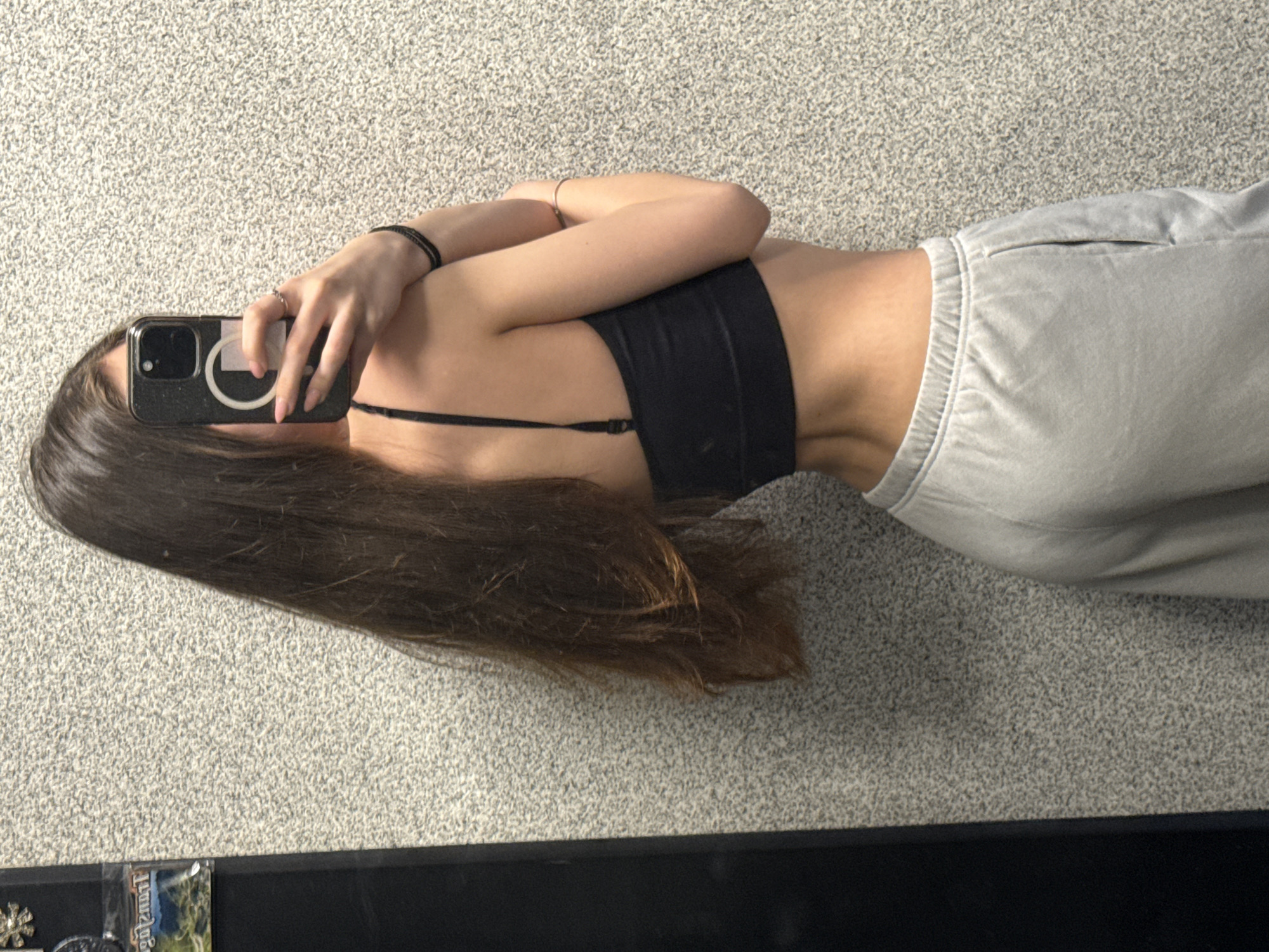 A mirror selfie captures a person, likely female, from the head down to just above the knees, against a light grey, speckled carpet background. The individual is positioned slightly to the right of the frame, with long, straight, dark brown hair flowing down her back and left side. Her face is obscured by a black smartphone, held up by her left hand, which also features a white circular ring holder on its back. She wears a black sports bra or bralette and light grey, elastic-waisted sweatpants.
A thin black bracelet adorns her left wrist, and a ring is visible on her left hand's ring finger. Her right arm is bent, resting across her midsection. The person's posture highlights a slim waist and a slight arch in her back. At the bottom of the image, a dark, flat surface, possibly a shelf or piece of furniture, is visible with a few small, indistinct items. One item partially displays the text "Jungle". The overall setting is an indoor space, likely a casual room with carpeted flooring, located in Sofia, Bulgaria. The image appears to be taken during the day, though specific time or weather cannot be determined.