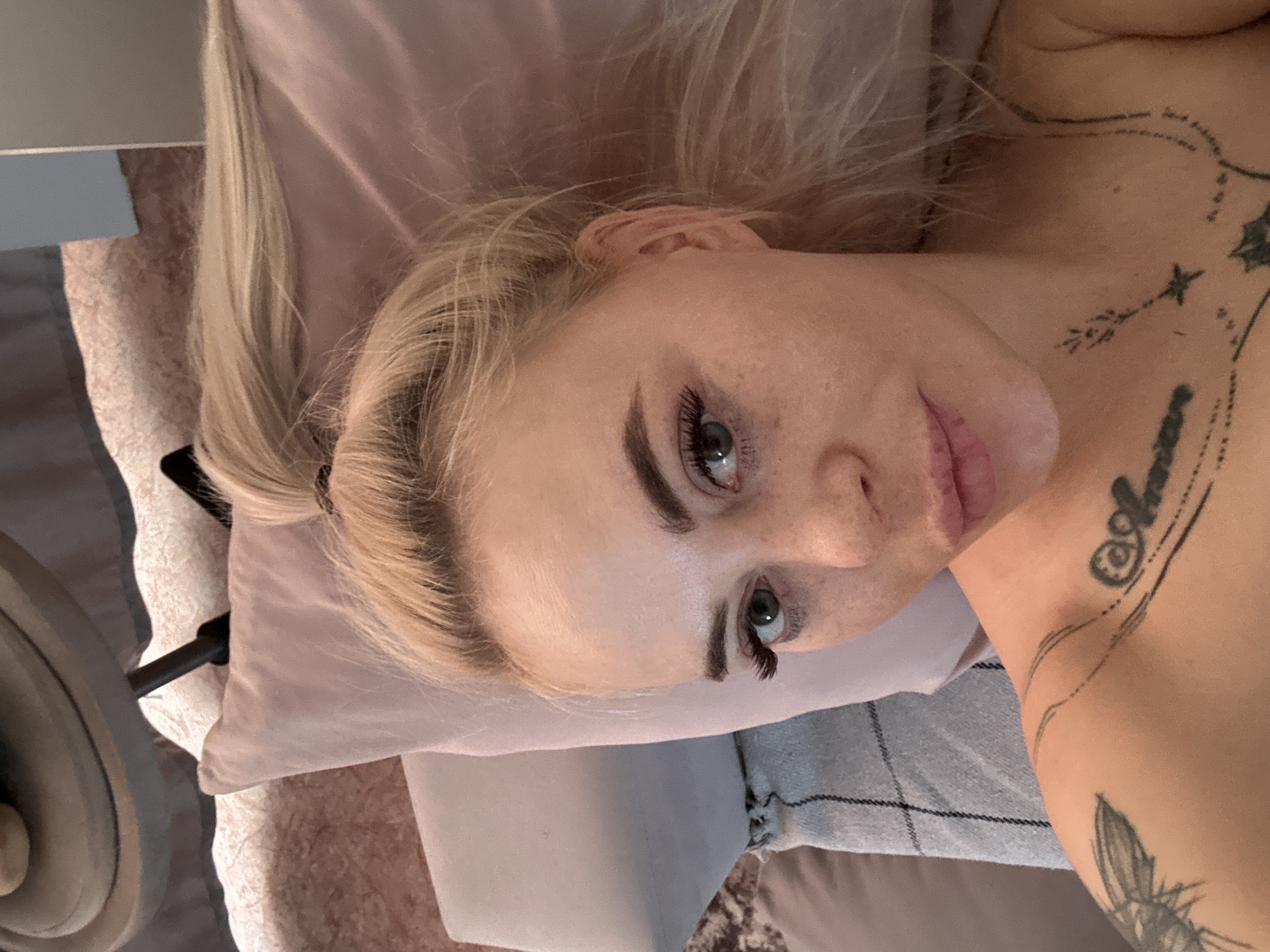 A person with light skin, blonde hair pulled back in a ponytail, and visible tattoos on their shoulder and upper chest is lying on what appears to be a bed or couch with pillows. The setting appears to be indoors, possibly a residential space. The city of Высокий, Russia, is not discernable in this image.