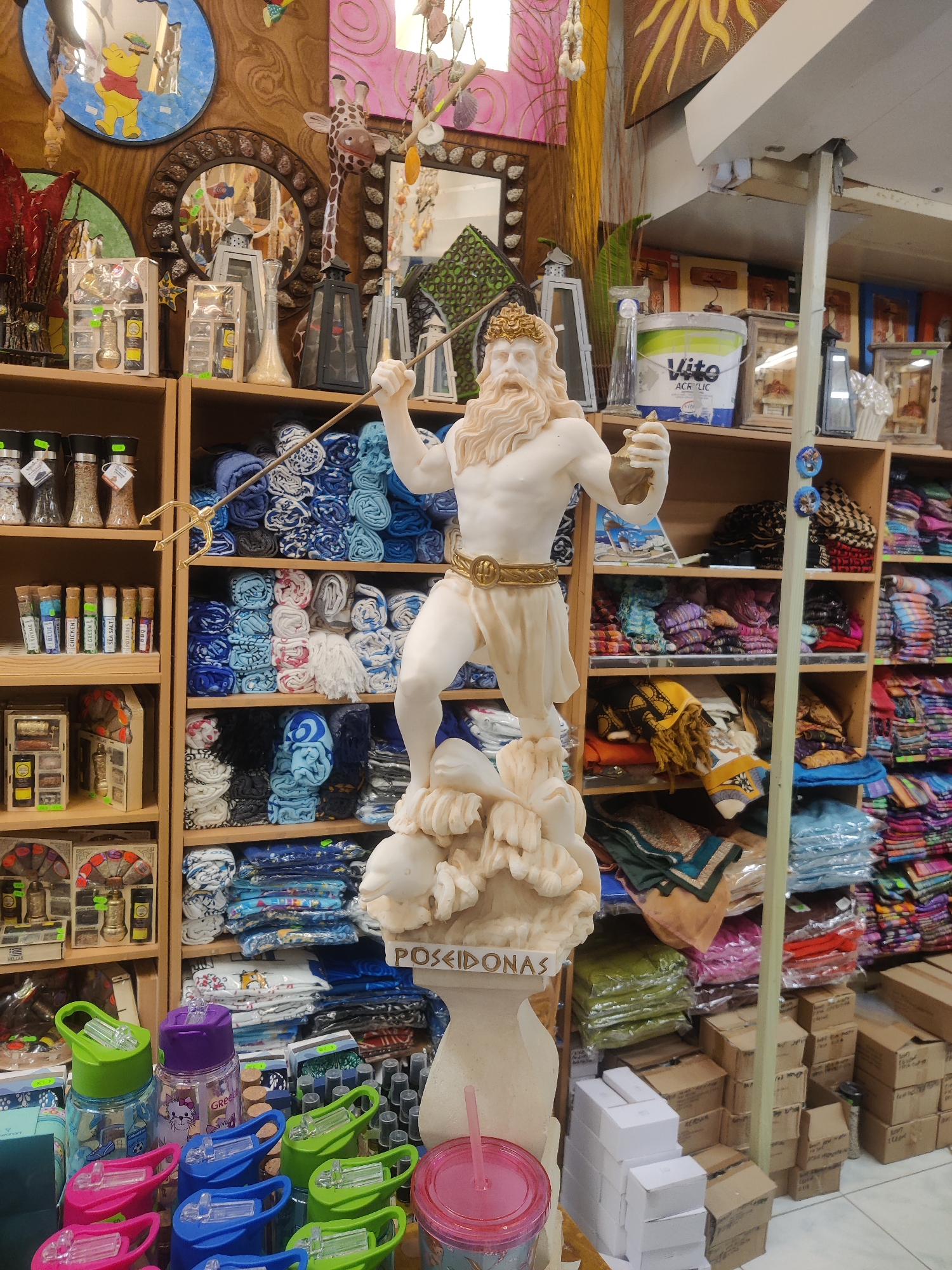The image depicts a retail display, likely in a souvenir or gift shop, featuring a statue of Poseidon prominently in the foreground. The statue is a pale, off-white color and stands on a white base that resembles waves or rocks. Poseidon is depicted with a beard and crown, holding a golden trident. Behind the statue, shelves are filled with various items, including rolled-up blue and white towels or fabrics, as well as colorful textiles. To the left of the statue, on lower shelves, are various small bottles and boxes containing spices or seasonings, and decorative items such as mirrors and lanterns. In the lower foreground, several brightly colored water bottles with plastic lids are visible. The overall impression is that of a crowded marketplace or a shop specializing in decorative and functional goods. The location is specified as Katerini Municipality, Greece, which suggests a tourist-oriented setting. The scene is well-lit, highlighting the details of the merchandise. There are no people visible in the image.