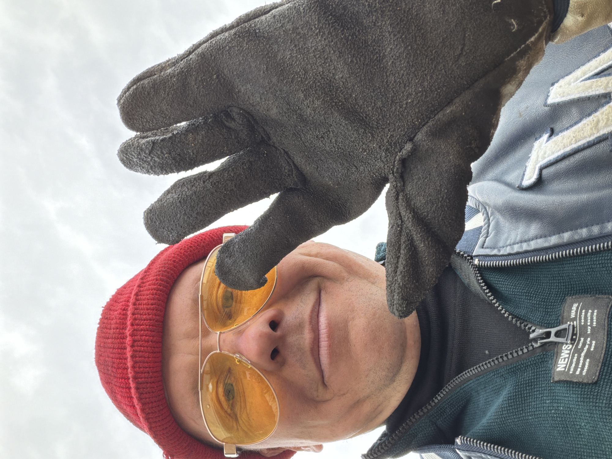 A man wearing a red beanie and yellow-tinted aviator sunglasses is viewed from a low angle. He is wearing a dark jacket with white lettering and a grey glove covering part of his face. The background is a cloudy sky. The location is unknown and there is no evidence to suggest it is Chaikovsky, Russia.