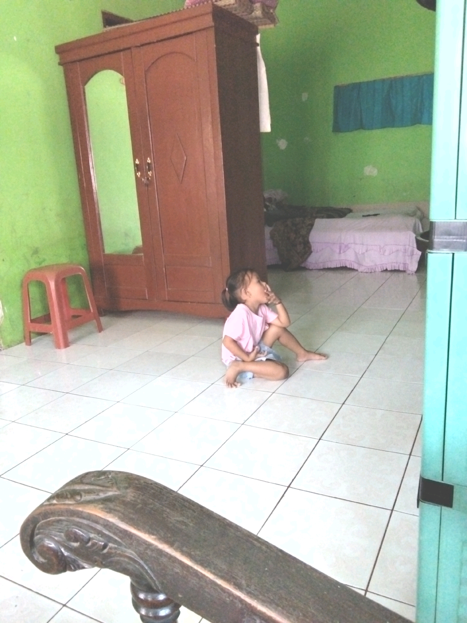 In a room with pale green walls and a tiled floor, a young girl in a pink t-shirt and blue shorts sits cross-legged. She is looking to her right with her left hand touching her chin. To her left, a red plastic stool stands beside a dark wooden wardrobe with a mirrored door. In the background, a bed is covered with a white bedspread and a patterned throw. A blue curtain hangs on the wall above the bed. A dark wooden armrest from a piece of furniture is in the foreground, partially obscuring the view. The scene appears to be in a residential setting in Rahong, Indonesia.