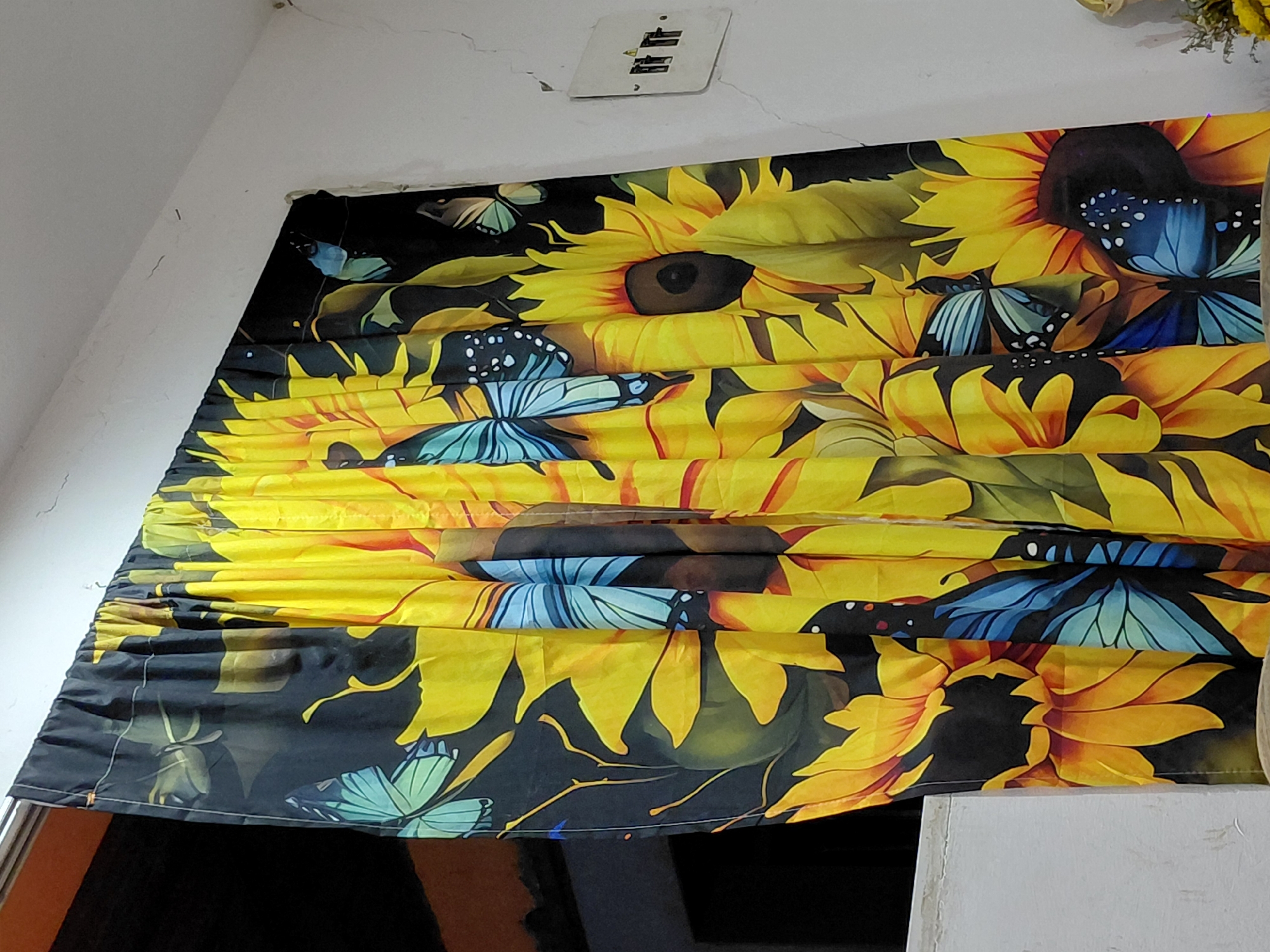 The image shows a window covering featuring a print of sunflowers and butterflies in shades of yellow, black, and blue. The covering is partially drawn, creating folds and wrinkles. The location is a room with white walls. The location within Santa Ana, Venezuela cannot be determined based on the provided image.