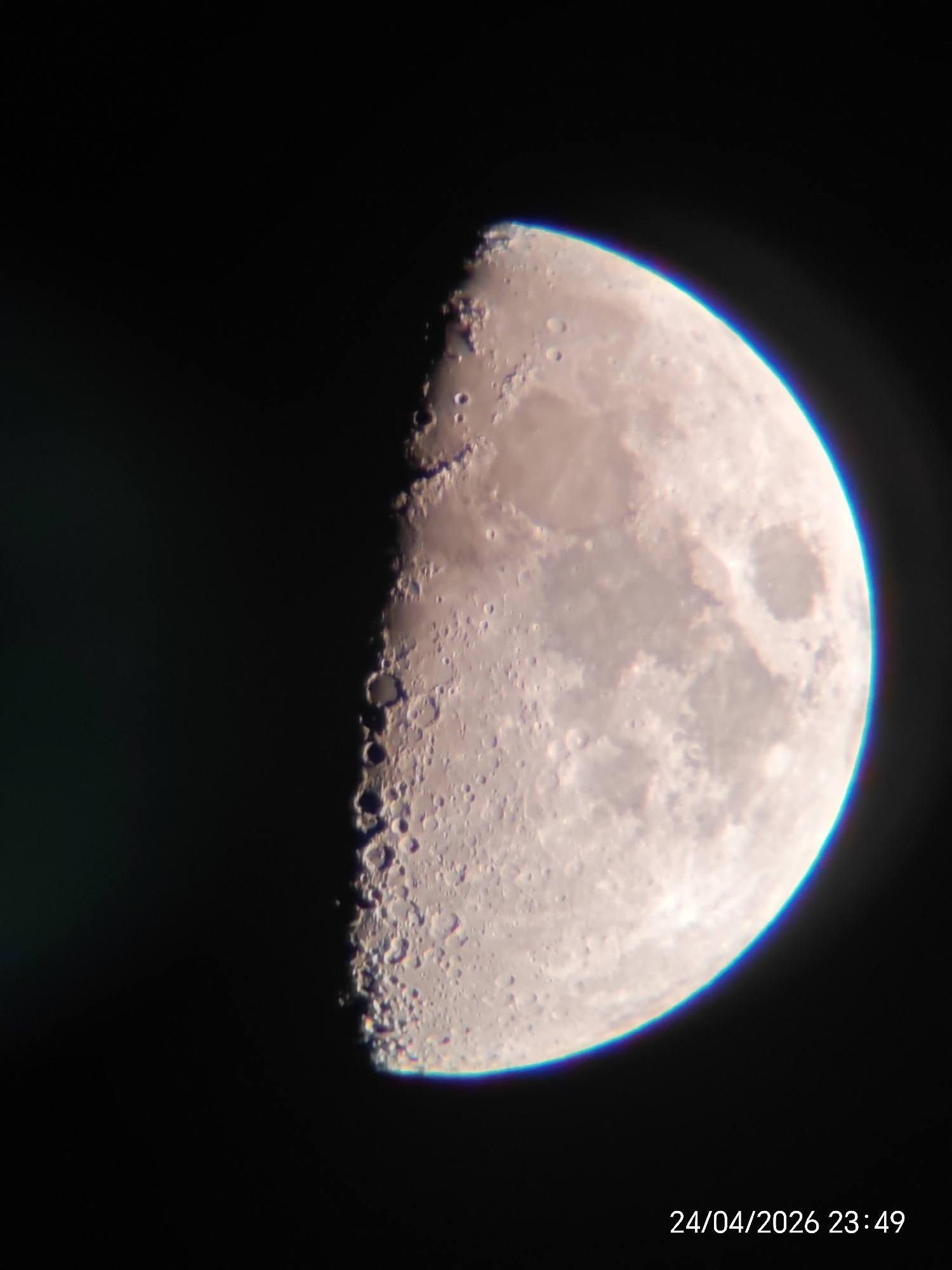 The image presents a close-up, detailed photograph of a half-illuminated Moon set against a deep black background, characteristic of a night sky or space. The Moon, appearing to be in its first or third quarter phase, fills the right half of the frame. Its surface is rich with visible features, including numerous impact craters of varying sizes, which are particularly prominent along the terminator – the distinct line separating the illuminated and shadowed portions. Darker, relatively smoother areas, likely lunar maria, are also discernible across the brighter regions.
A notable optical detail is the presence of chromatic aberration, visible as a distinct blue and purple halo along the outer, brightly lit edge of the Moon, especially noticeable on the top and bottom right curves. The Moon is brightly lit, presumably by the sun, from the right side, creating sharp shadows within the craters and enhancing their relief along the terminator.
In the bottom right corner of the image, white text displays the timestamp "24/04/2026 23:49," indicating the precise date and time the photograph was captured. The overall impression is that of a clear, unclouded night, enabling a detailed astronomical observation of the Moon.