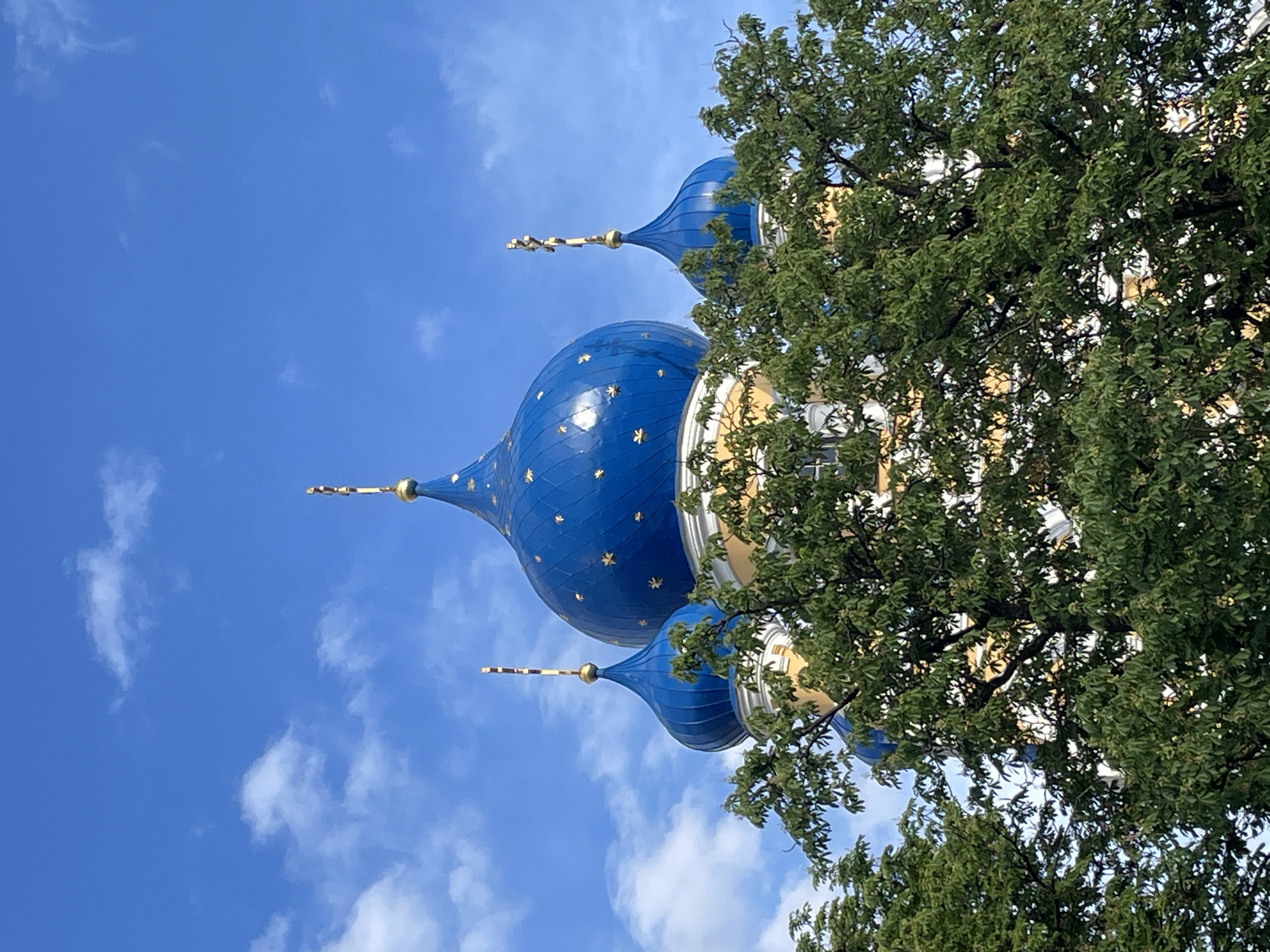The image displays a partial view of the Saints Peter and Paul Cathedral in Brest, Belarus, under a bright blue sky featuring scattered white cumulus clouds. Three blue, onion-shaped domes are prominently featured, with the largest central dome flanked by two smaller ones. Each dome is adorned with numerous small golden stars and culminates in a golden finial topped with an ornate golden Orthodox cross. Portions of the cathedral's structure visible beneath the domes are painted light yellow with white architectural accents. The lower section of the building is largely obscured by dense, green foliage from deciduous trees in the foreground. No individuals or specific activities are present within the frame.
