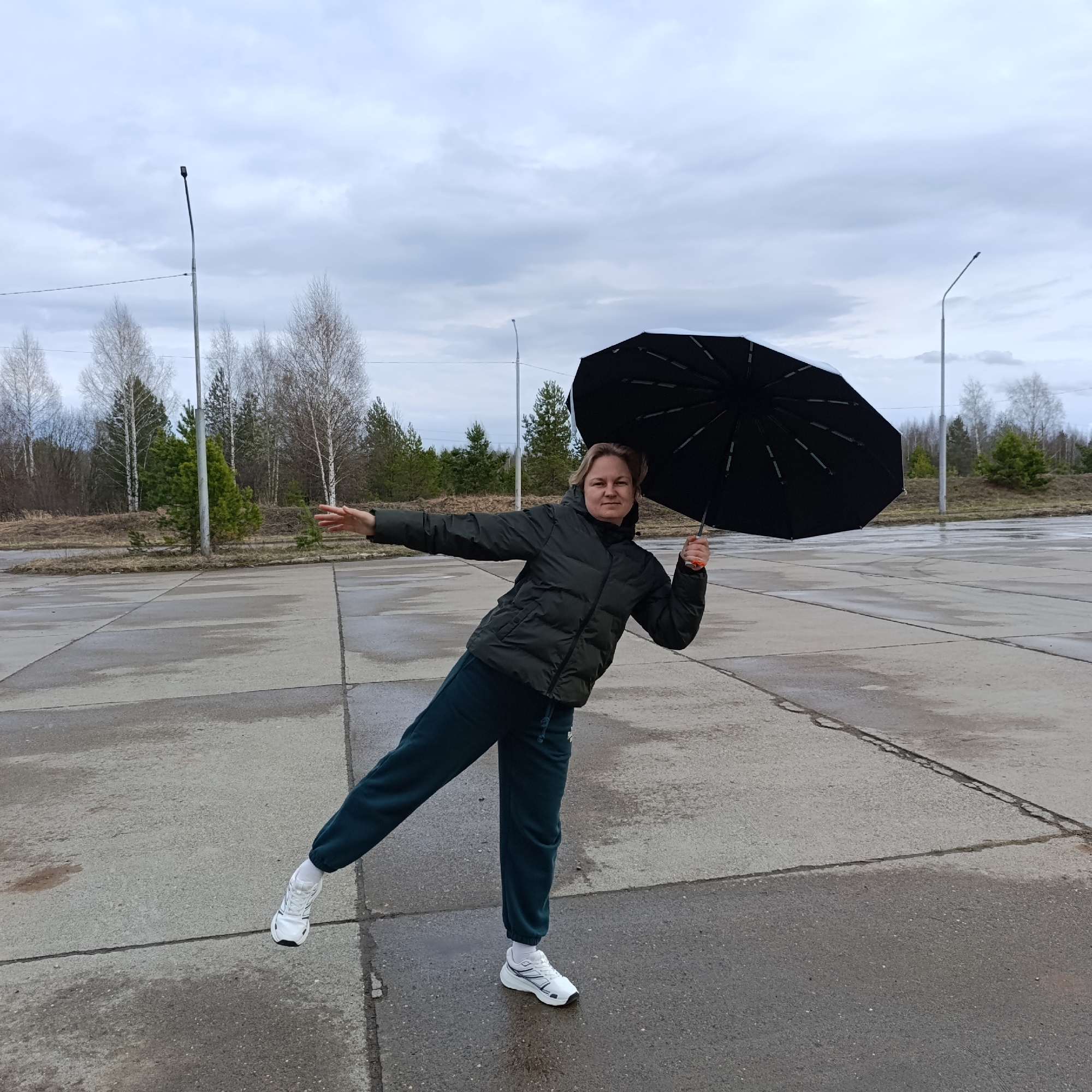 A woman stands on a large concrete surface with trees in the background. She is holding a black umbrella in her right hand, which is raised. Her left arm is extended to the side. She wears a dark green puffer jacket and dark green sweatpants, with white sneakers. Her left leg is raised, giving the impression of balancing. The sky is overcast, and the ground appears damp. Two light poles are visible in the background, along with bare trees and evergreen trees. The location is Markovskii, Russia, based on the information provided.