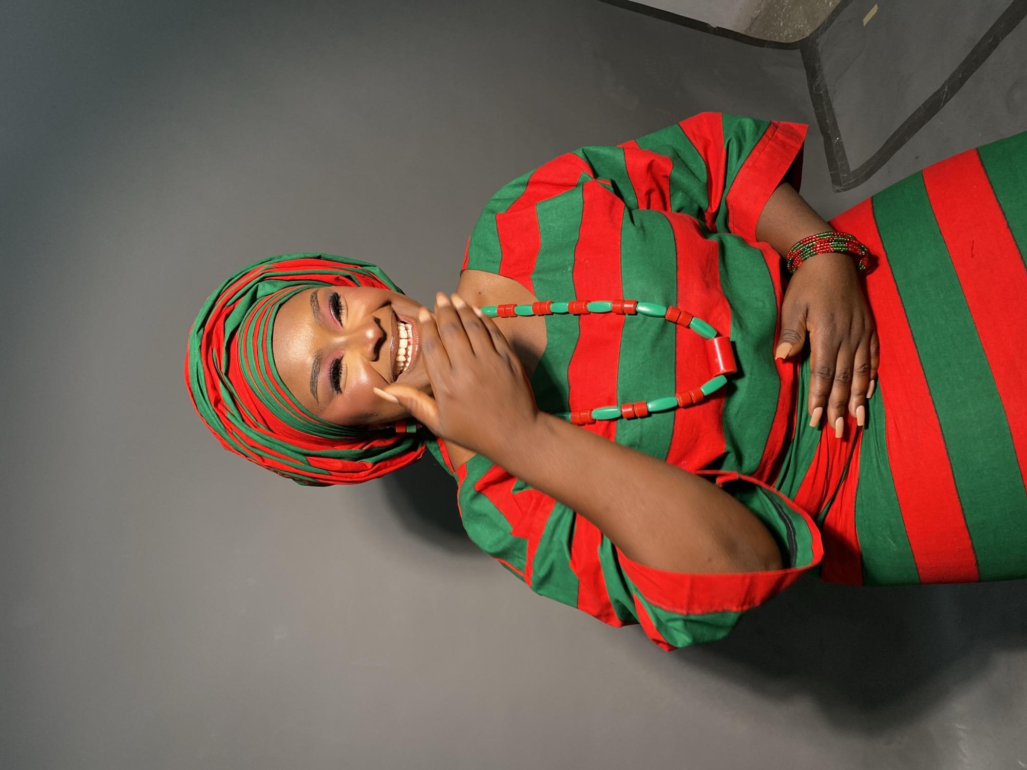A woman smiles while posed indoors in what appears to be a studio. She is wearing a red and green striped dress and headwrap, a necklace of red and green beads, and a red and green beaded bracelet. She has her hand near her face. The background is a gray wall. Location in Gura Top, Nigeria, is not verified.