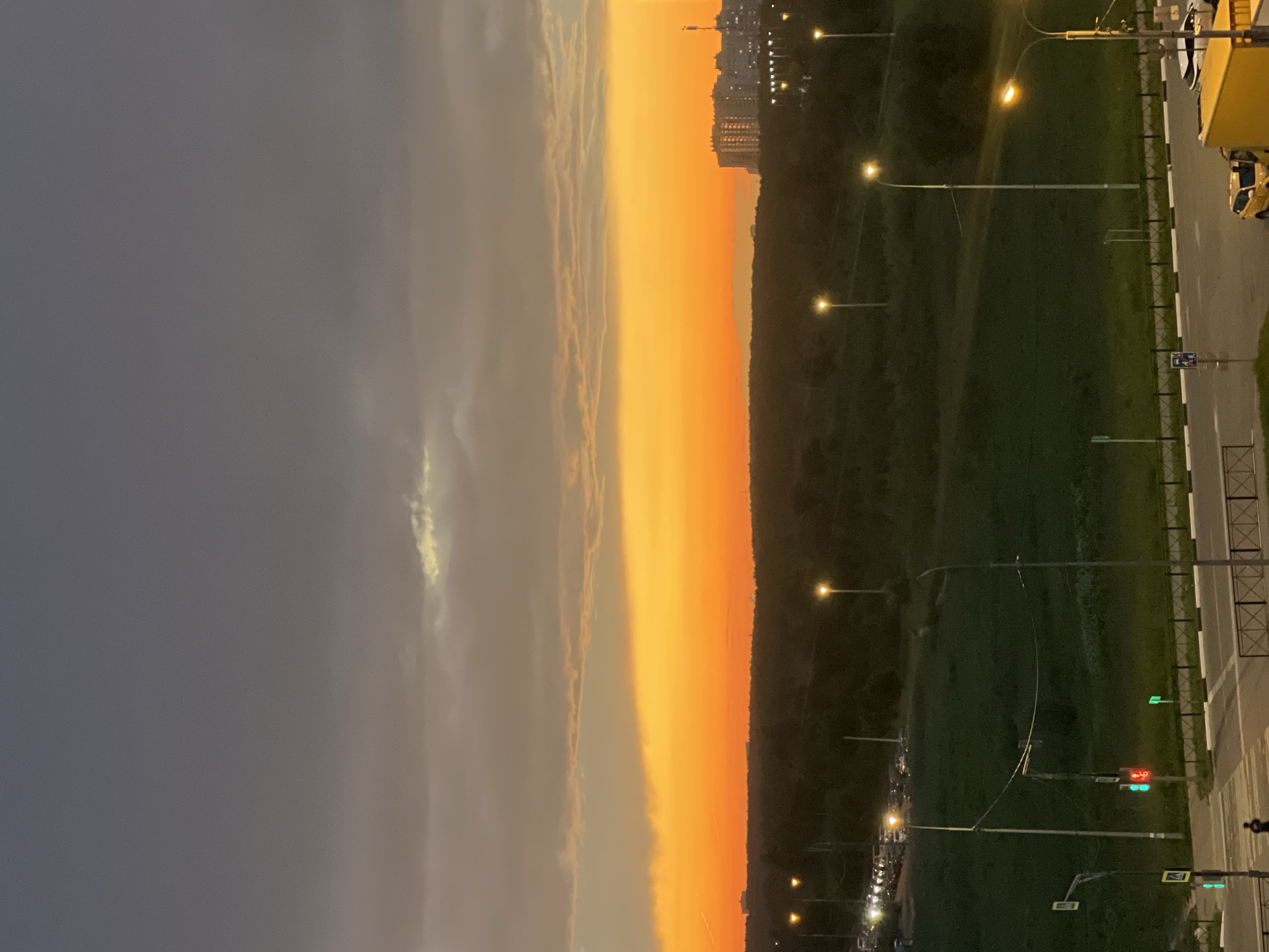 A vertical photograph captures a dusky sky transitioning from a dark gray on the left to a vibrant orange sunset on the right. Thin, wispy clouds are visible within the gray section, catching some of the sunset's light.
The right side of the image depicts a scene with a road, a grassy area, and streetlights. A line of trees separates the grassy area from a distant cityscape featuring tall buildings illuminated by lights. Several streetlights are visible, some with their lights on, illuminating the scene. Along the road, there are white and black striped barriers. A yellow truck is parked on the right edge of the frame.
A traffic light displaying red and green signals is visible, along with pedestrian crossing signs. The overall impression is an urban or suburban setting at dusk or early evening, as indicated by the sunset colors and the illuminated streetlights. There is no discernible text in the image. The mood is serene, with the dramatic sunset contrasting with the approaching darkness.