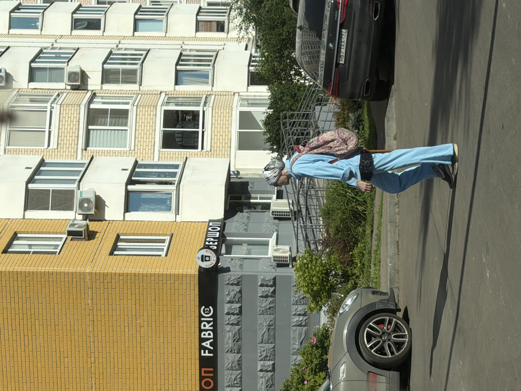 A woman walks across a street in front of a multi-story residential building in Moscow, Russia. The building has a facade of yellow brick on the lower floors and cream-colored siding on the upper floors. Several windows are visible, some with air conditioning units mounted beneath them. A store with the name "OP FABRIC" is located on the first floor, next to a coffee shop called "BEPШON". The woman is wearing a light blue tracksuit and a headscarf. She has a pink and white patterned backpack on her back. She is walking from left to right across the frame. A grey car is parked on the left side of the street, and a black BMW is parked on the right side. The ground is asphalt, and there is a sidewalk with greenery and bushes. The lighting suggests it is a sunny day.
