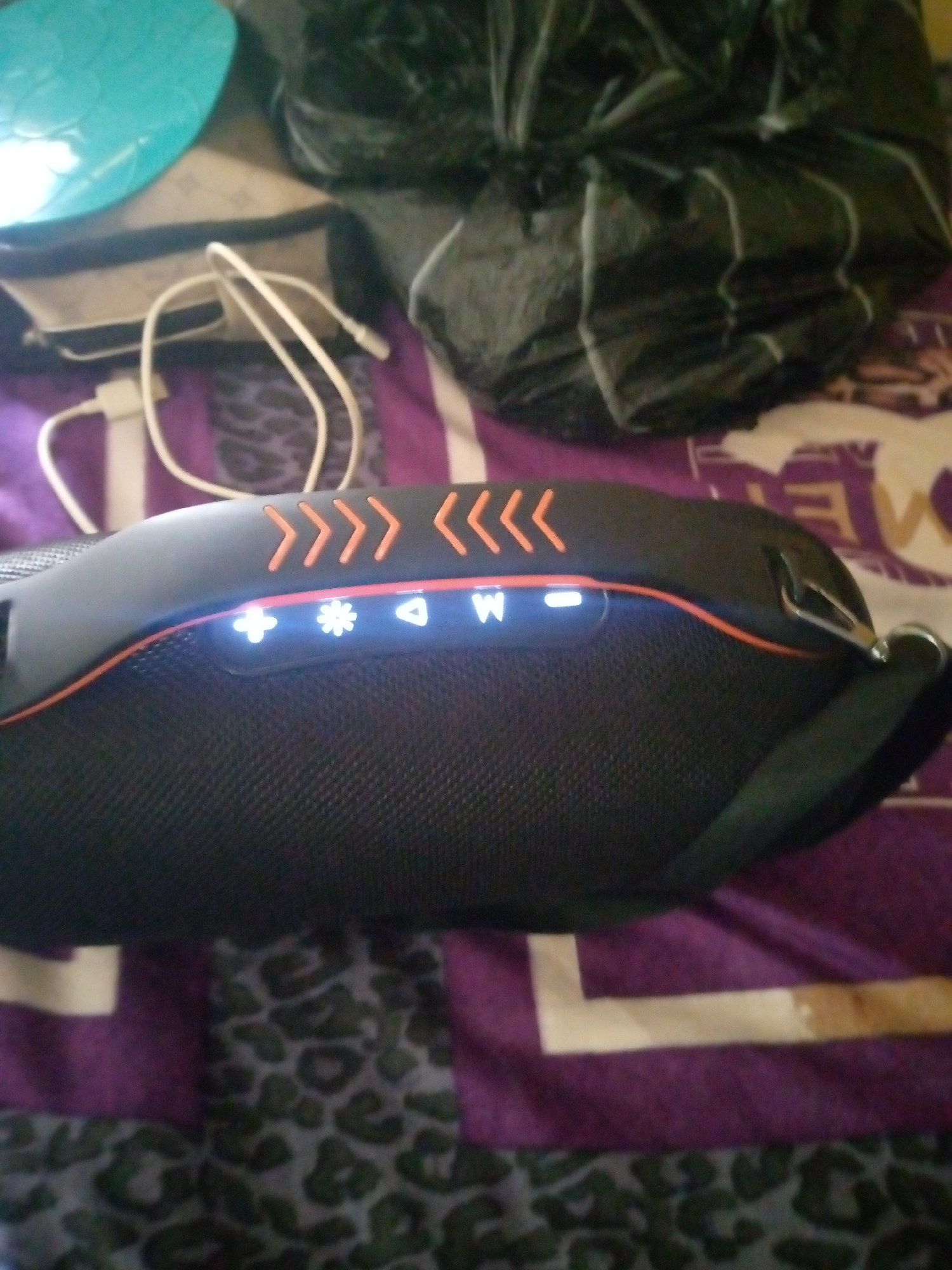 The image shows a close-up of a black portable speaker with orange accents. Several illuminated symbols are visible on the speaker's surface, including a plus sign, a snowflake, a triangle, and the letter "W". A white USB cable is connected to the speaker and lies loosely to the left. The speaker is placed on a surface with a purple and leopard print pattern. In the background, out of focus, are a bright blue object and a dark, crumpled material. The scene appears to be indoors, on a bed or sofa, in Jalingo, Nigeria.