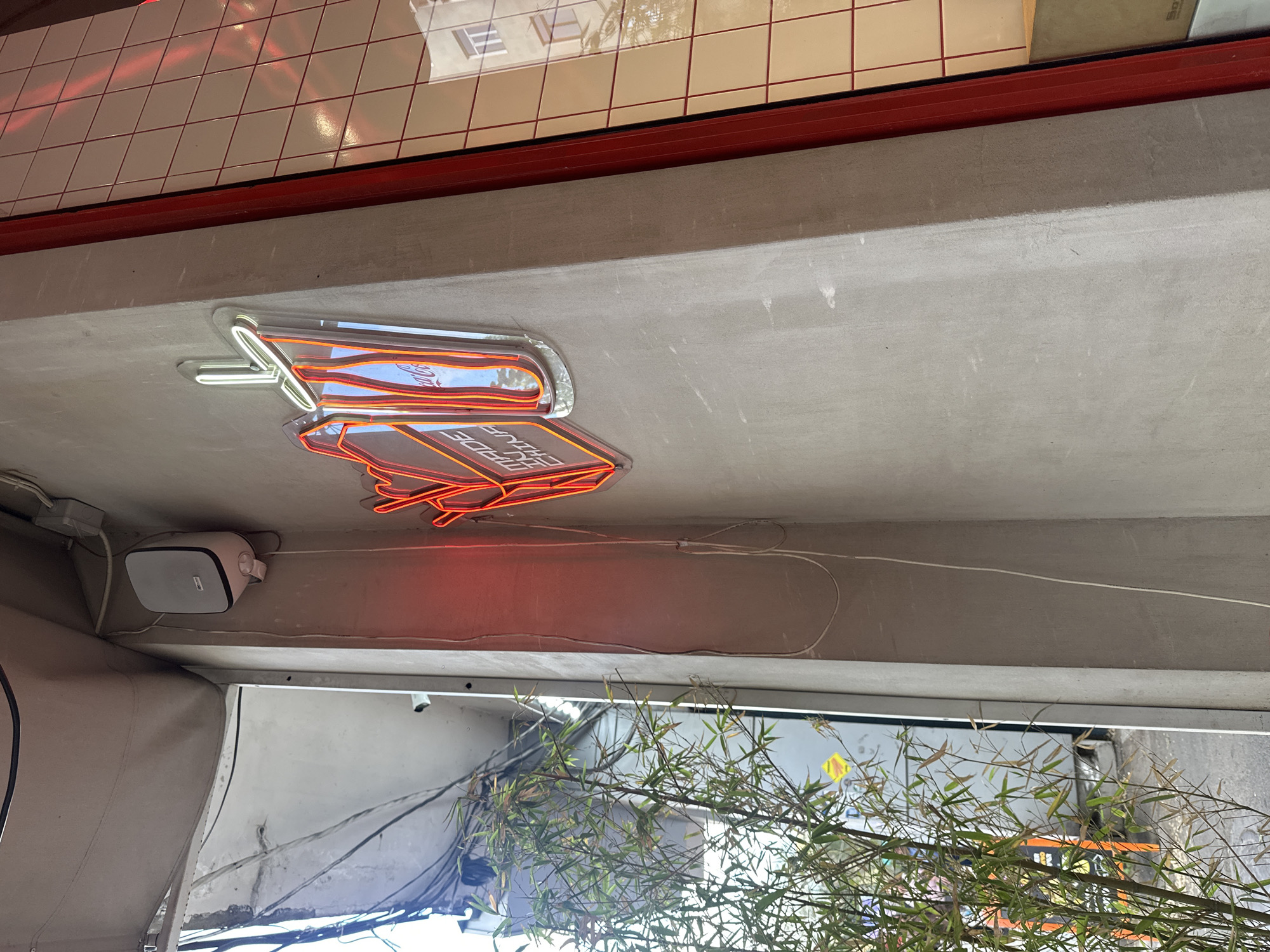 The photograph shows a partial, upward-angled view of an outdoor setting in Yerevan, Armenia, focusing on architectural and decorative elements. A section of a ceiling or overhang is prominently featured, with a gray, textured surface. Mounted on the ceiling is a white, box-shaped object, possibly a speaker.
A neon sign is attached to the ceiling. It displays the words "TRADE EHOUSE" in capital letters. The sign is outlined in white neon tubing.
Below the ceiling, a dense arrangement of bamboo or similar plant foliage obscures some of the view. Through the foliage, glimpses of the city street can be seen. The background shows building structures and wiring.
Above the ceiling area, a narrow portion of a wall is visible. It is covered in a grid pattern of tiles with red grout lines. The overall impression is that of an urban outdoor space, possibly a storefront or restaurant patio, decorated with modern elements.