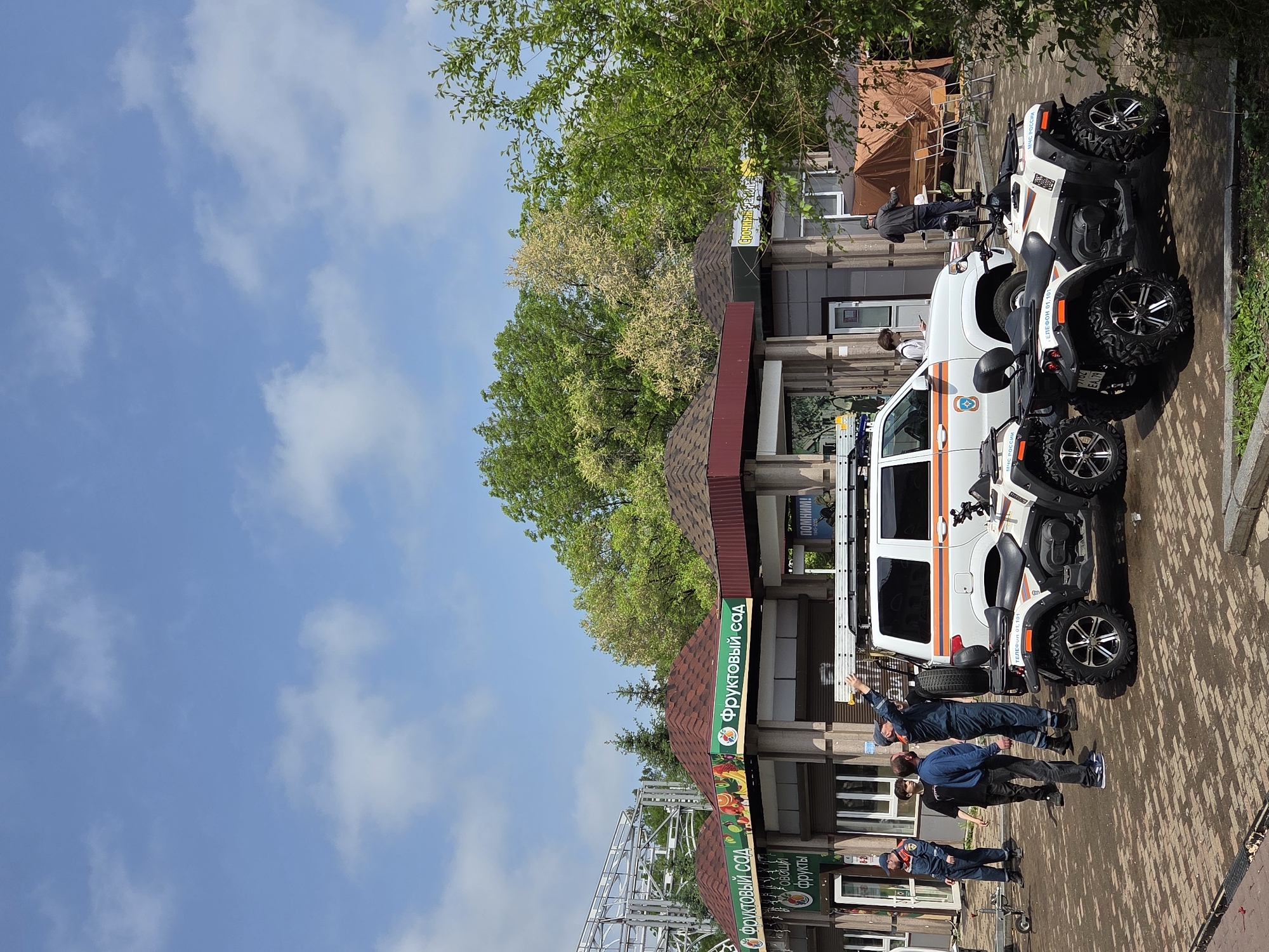 The image depicts a scene in Birobidzhan, Russia. A white emergency vehicle with orange stripes and the insignia of the Ministry of Emergency Situations (МЧС России) is parked next to a building with a sign reading "ФРУКТОВЫЙ САД" ("Fruit Garden") in Cyrillic. Three people wearing blue uniforms are standing near the vehicle. Two ATVs are parked behind the vehicle. The surrounding environment includes trees and a clear, blue sky with scattered clouds. The building appears to be a shop or business.