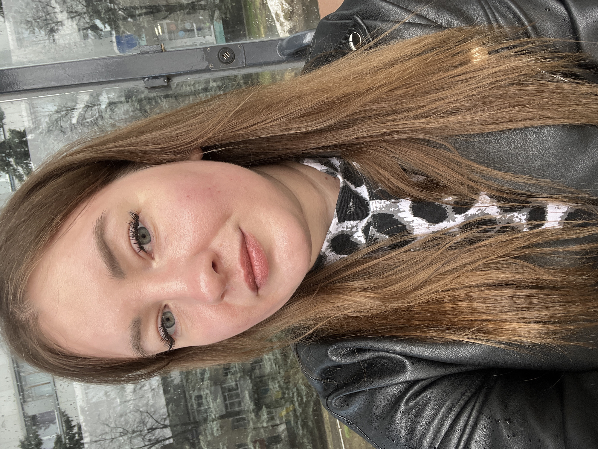 A close-up, angled selfie shows a light-skinned woman with long, light brown hair, green eyes, and pink lips, wearing a black leather jacket over a black and white patterned top. The background appears to be a window with blurred details possibly showing a street scene in Chaikovsky, Russia. Rain or condensation is visible on the window surface. A grey metal bar is visible in the top left corner of the image, possibly part of the window frame. The image is oriented vertically, as a phone selfie.