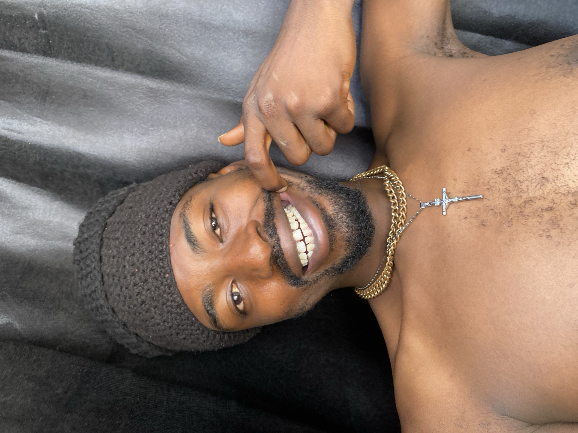 An adult man with dark skin is depicted lying on a dark, textured fabric surface, likely indoors in Barakin Akawo, Nigeria. He is positioned on his back or side, looking directly at the camera with a wide, expressive smile, showcasing his teeth. He wears a dark, possibly black, knitted beanie or cap and a gold chain necklace with a silver crucifix pendant.
The man's right hand is raised, with his index finger playfully pulling down the corner of his mouth, accentuating his smile. He has a beard and mustache. He is shirtless, revealing his upper torso with visible chest hair and some body hair in the armpit region. Several light marks or scars are visible on the back of his right hand. The lighting appears soft, illuminating his face and upper body. The overall impression is one of happiness and a playful demeanor. No text is visible in the image.